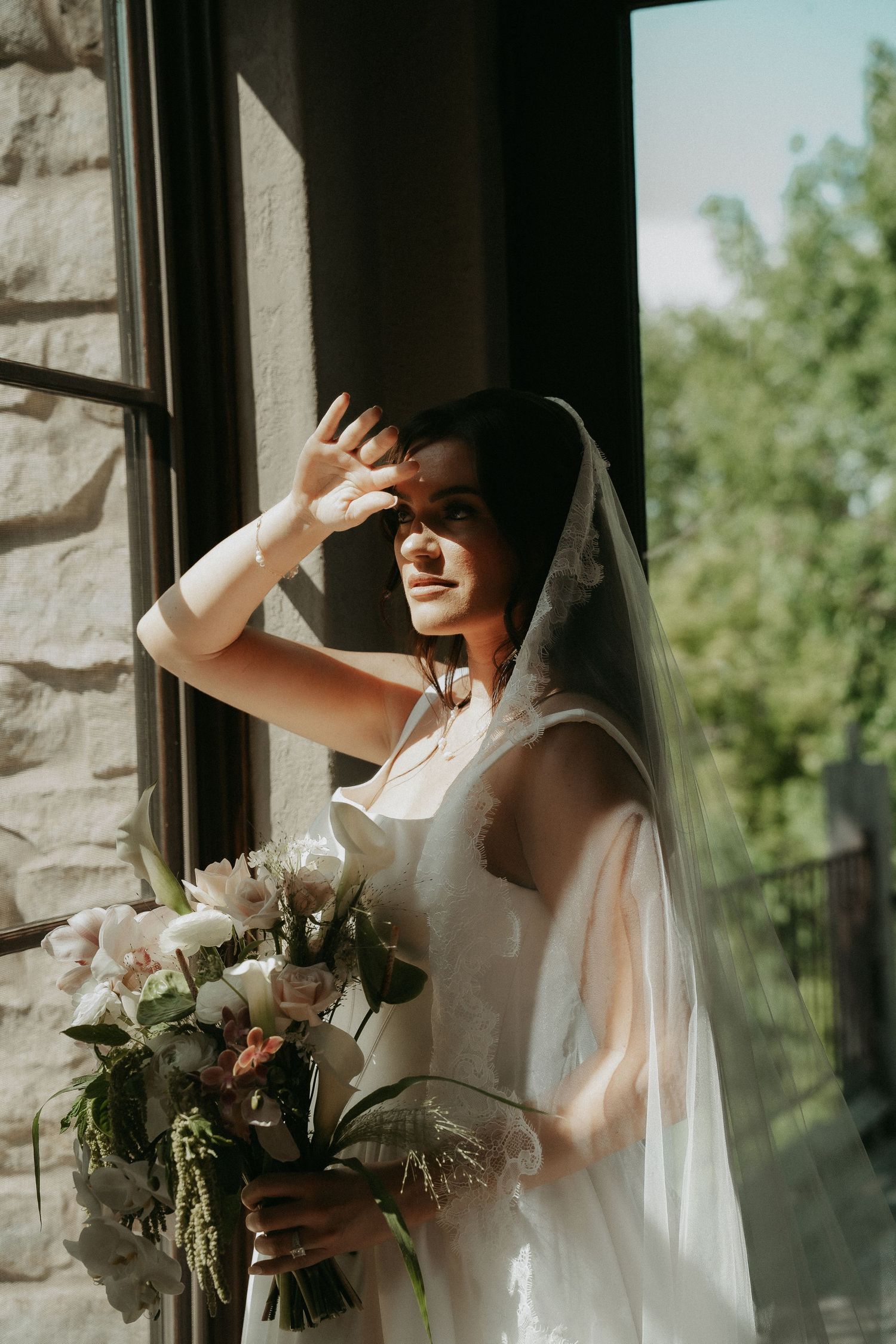 Bride in white gown and veil holds bouquet by sunlit window, shielding eyes from light.