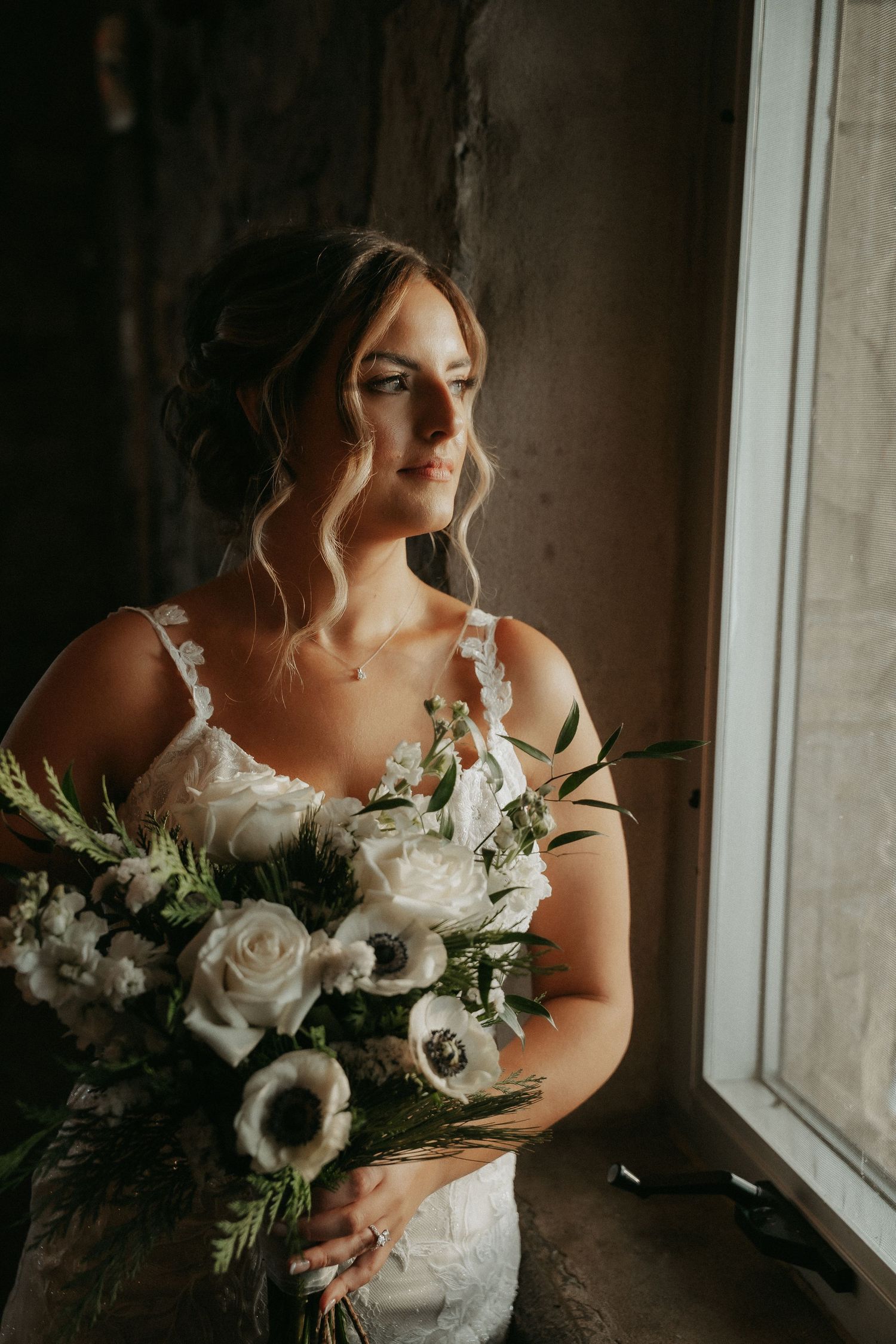 Bride in lace dress holds elegant white and green bouquet while standing near window in moody lighting.