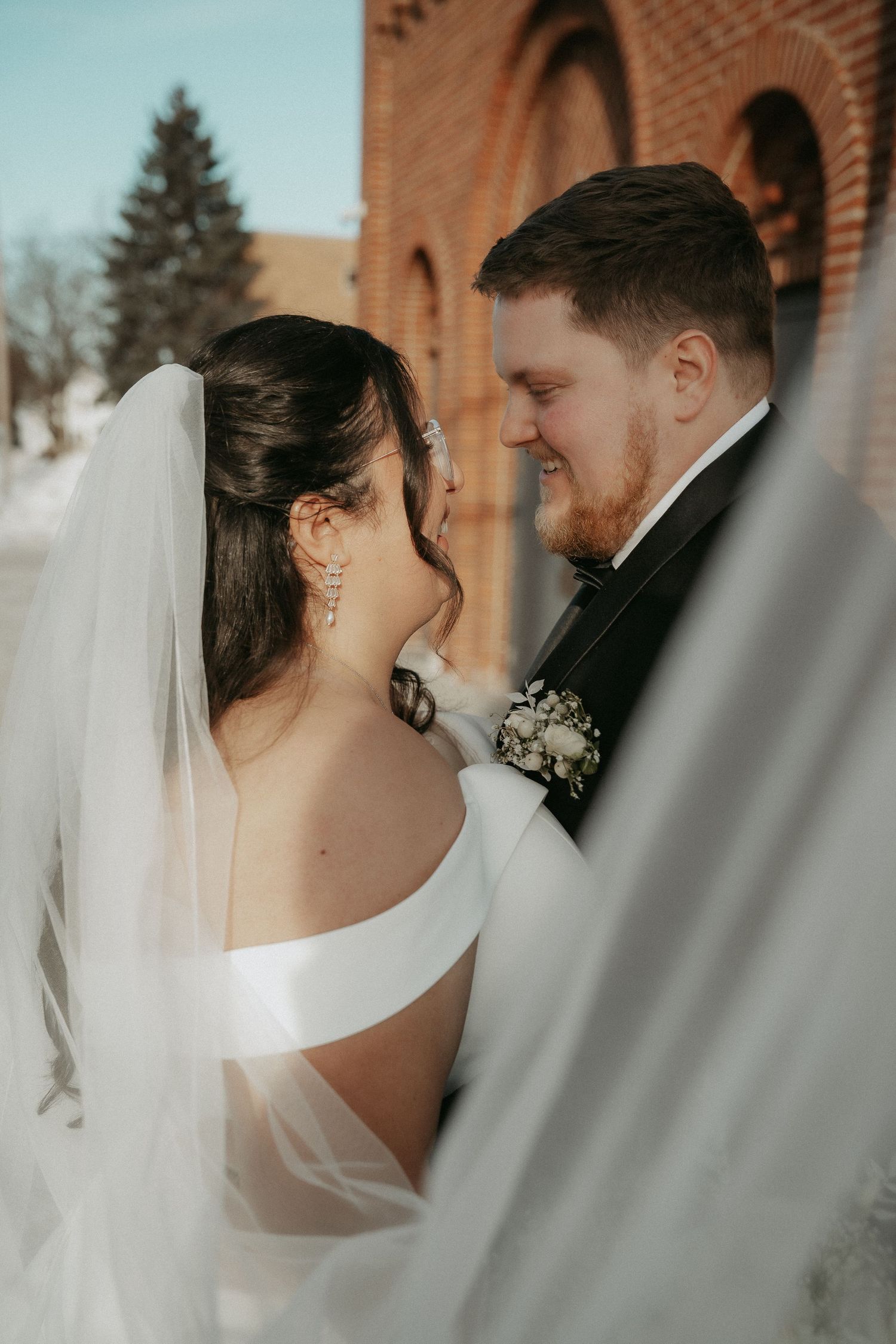 A tender wedding moment captured between a couple in front of a brick church with the bride wearing a flowing white veil.