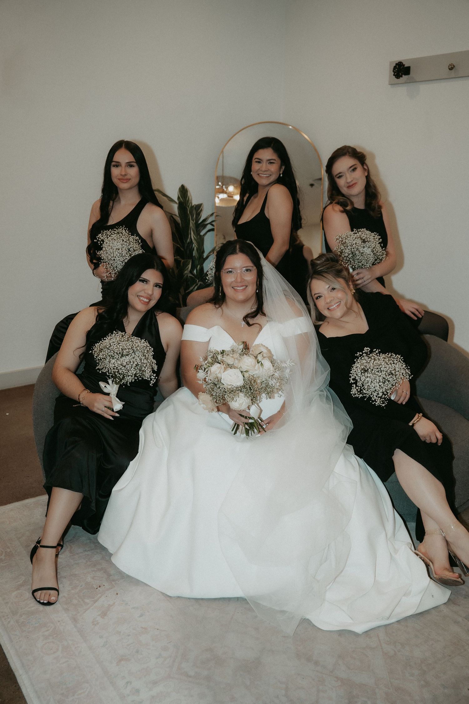 A bridal party poses together in black dresses and a white wedding gown holding bouquets in a minimalist setting.