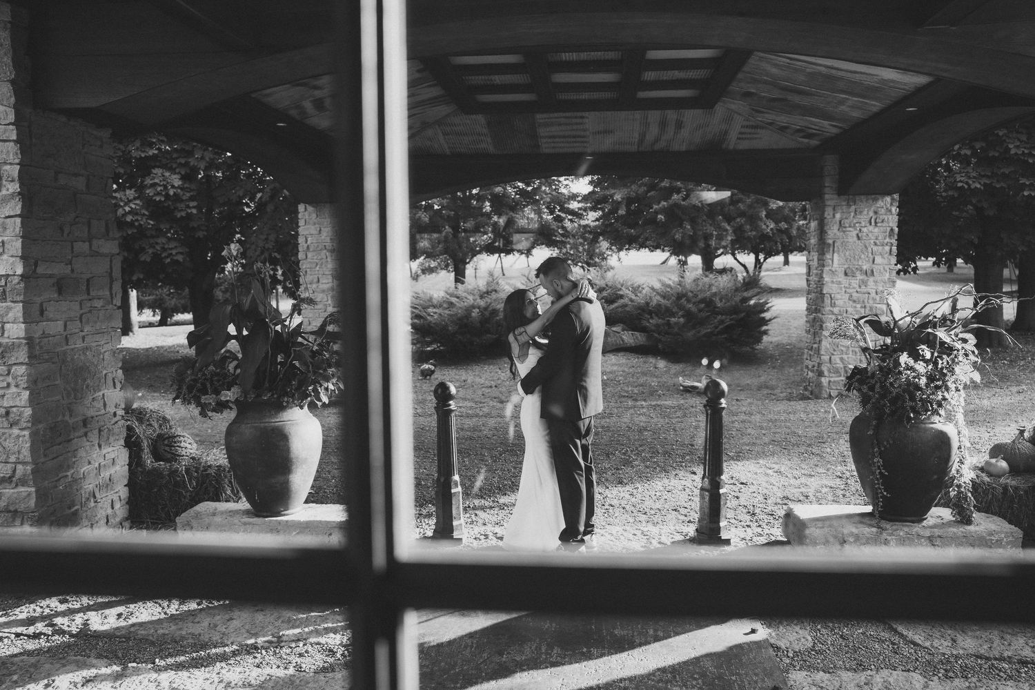 A black and white view through a window showing a romantic couple embracing under a covered outdoor patio.