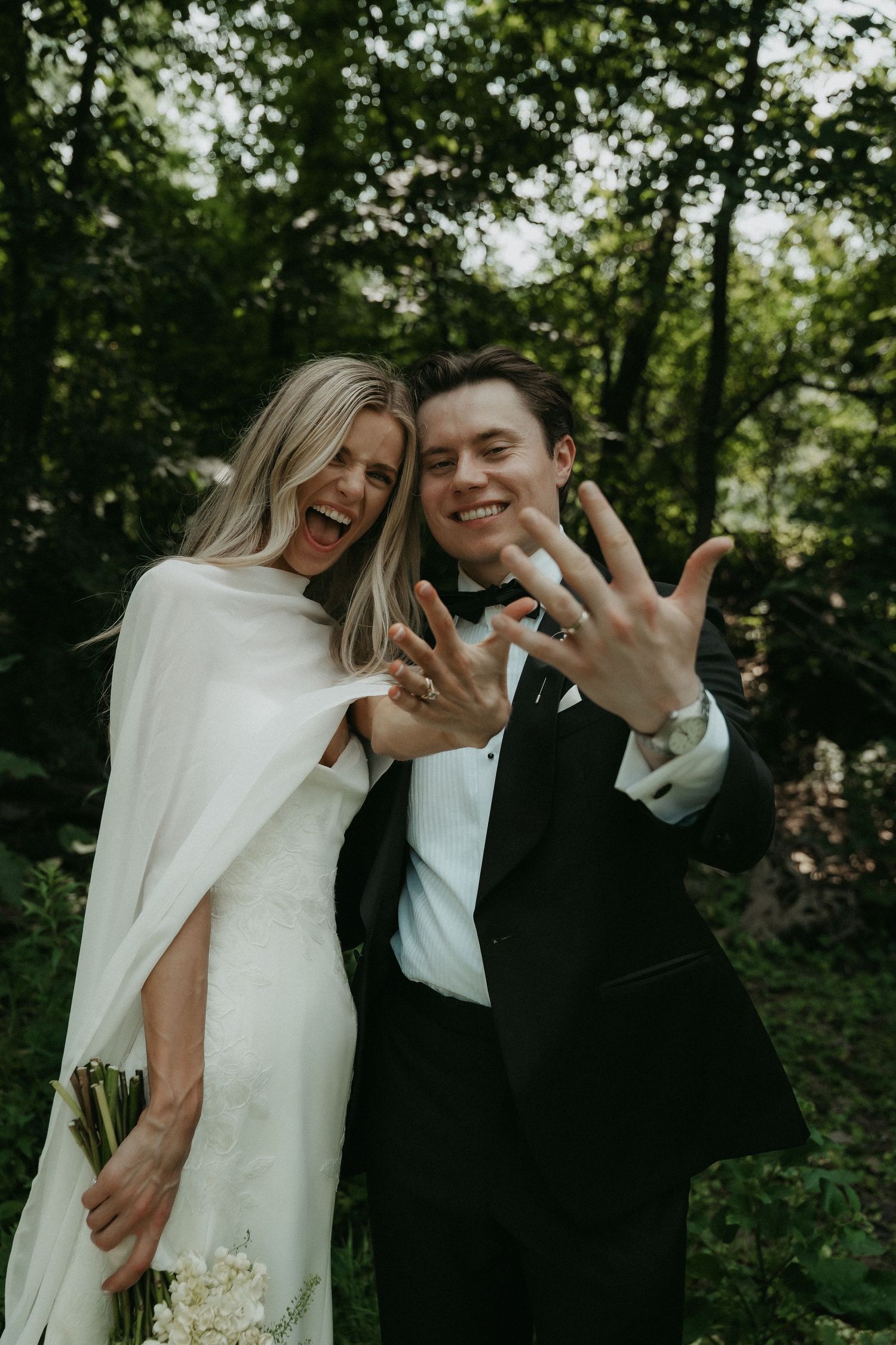 Joyful newlyweds showing off wedding rings in a lush green forest, bride in white cape gown holding bouquet.