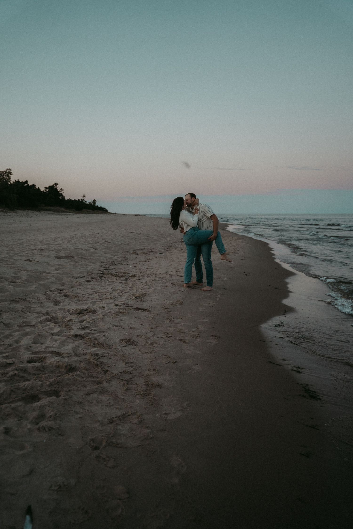 A couple walks along a misty beach shoreline at dusk with calm waves lapping at the sand.