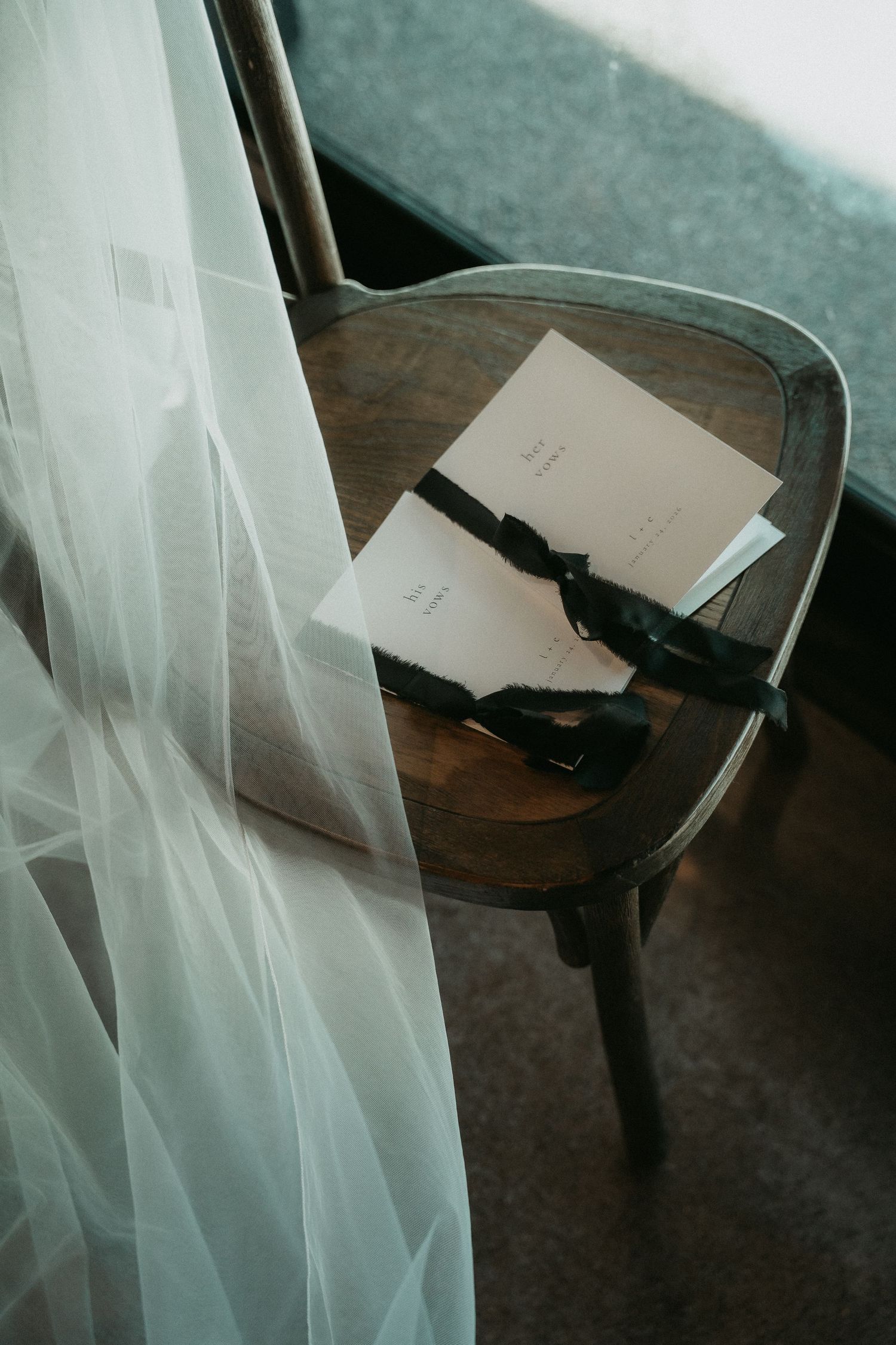 White sheer curtain blowing in breeze next to wooden chair with papers on dark floor in moody lighting.