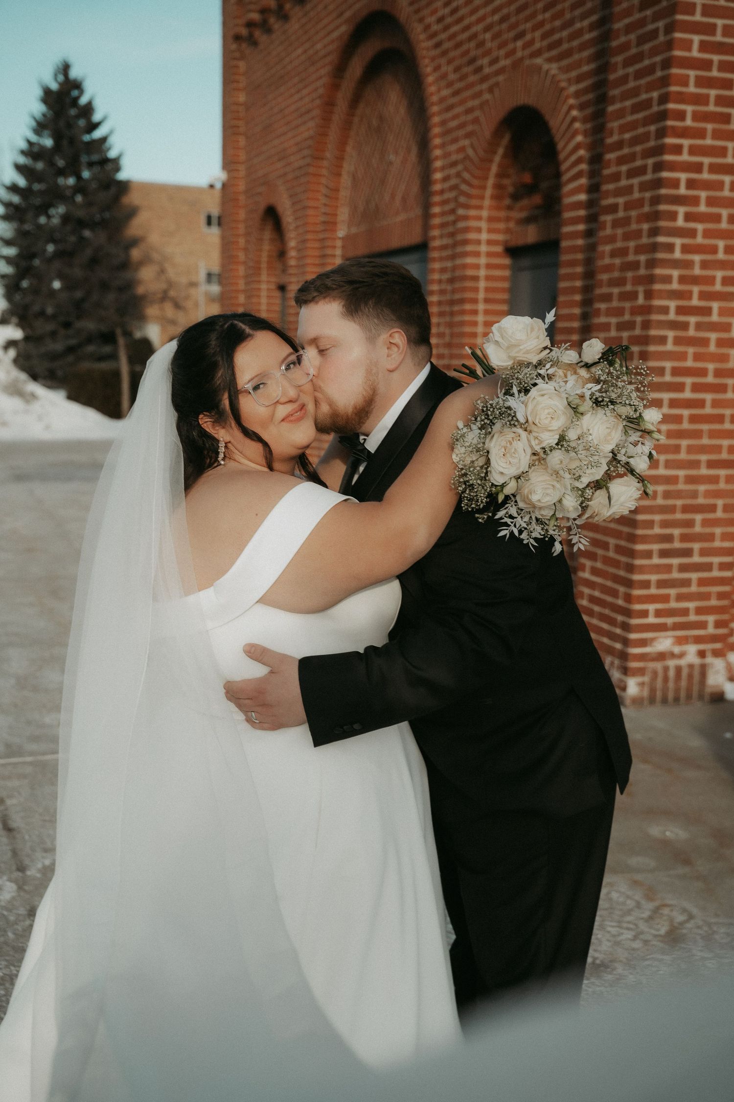 Romantic wedding couple shares intimate kisses in front of brick church arches during their winter ceremony.