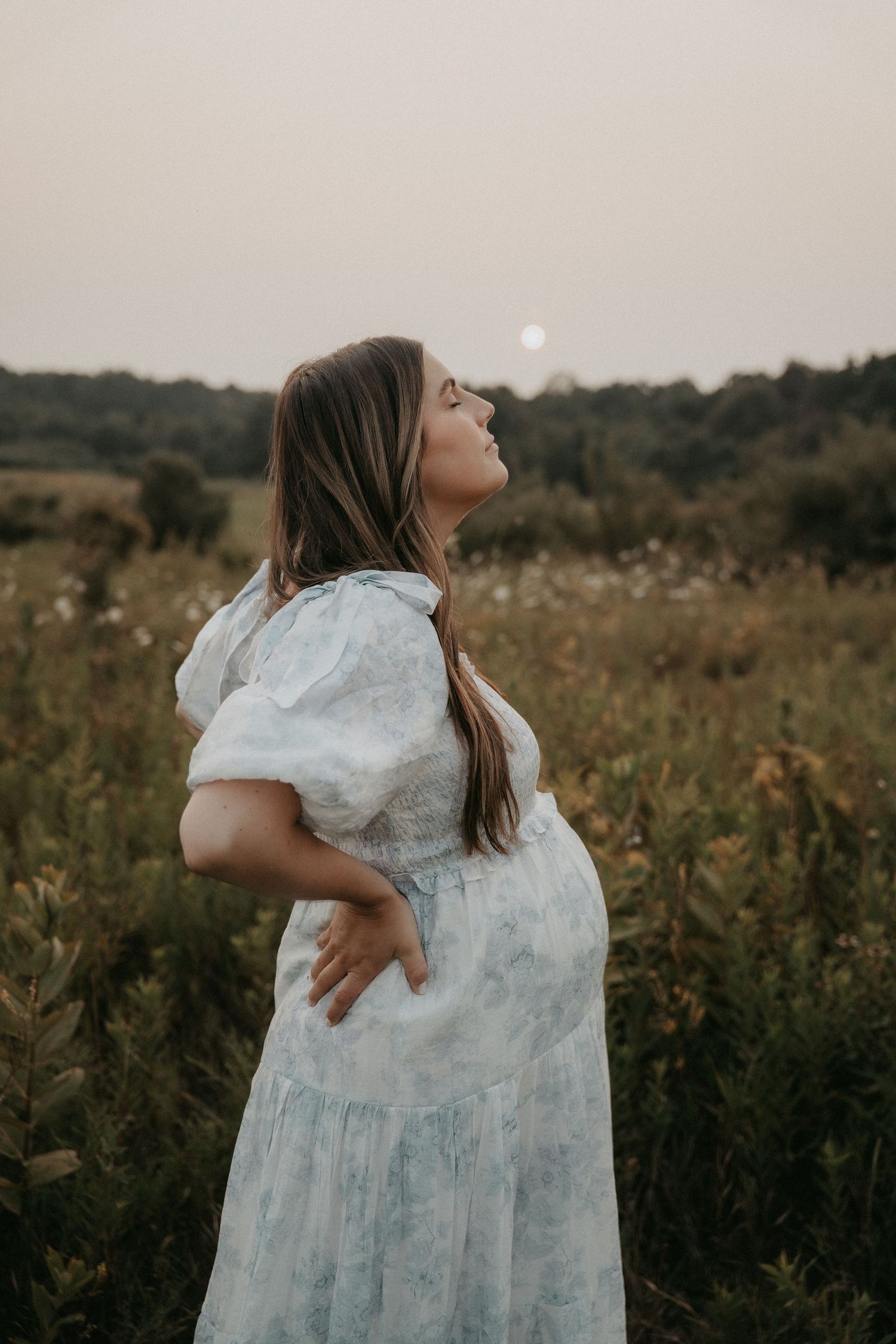 A person in a white flowy dress poses gracefully in a golden field at sunset.