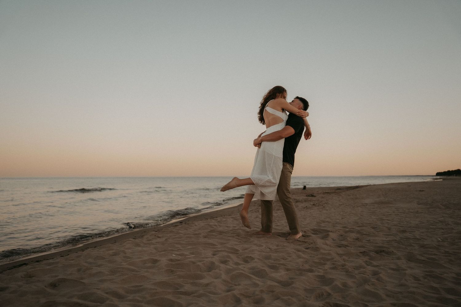 Two silhouettes embrace on a sandy beach at sunset with warm golden light illuminating the romantic scene.