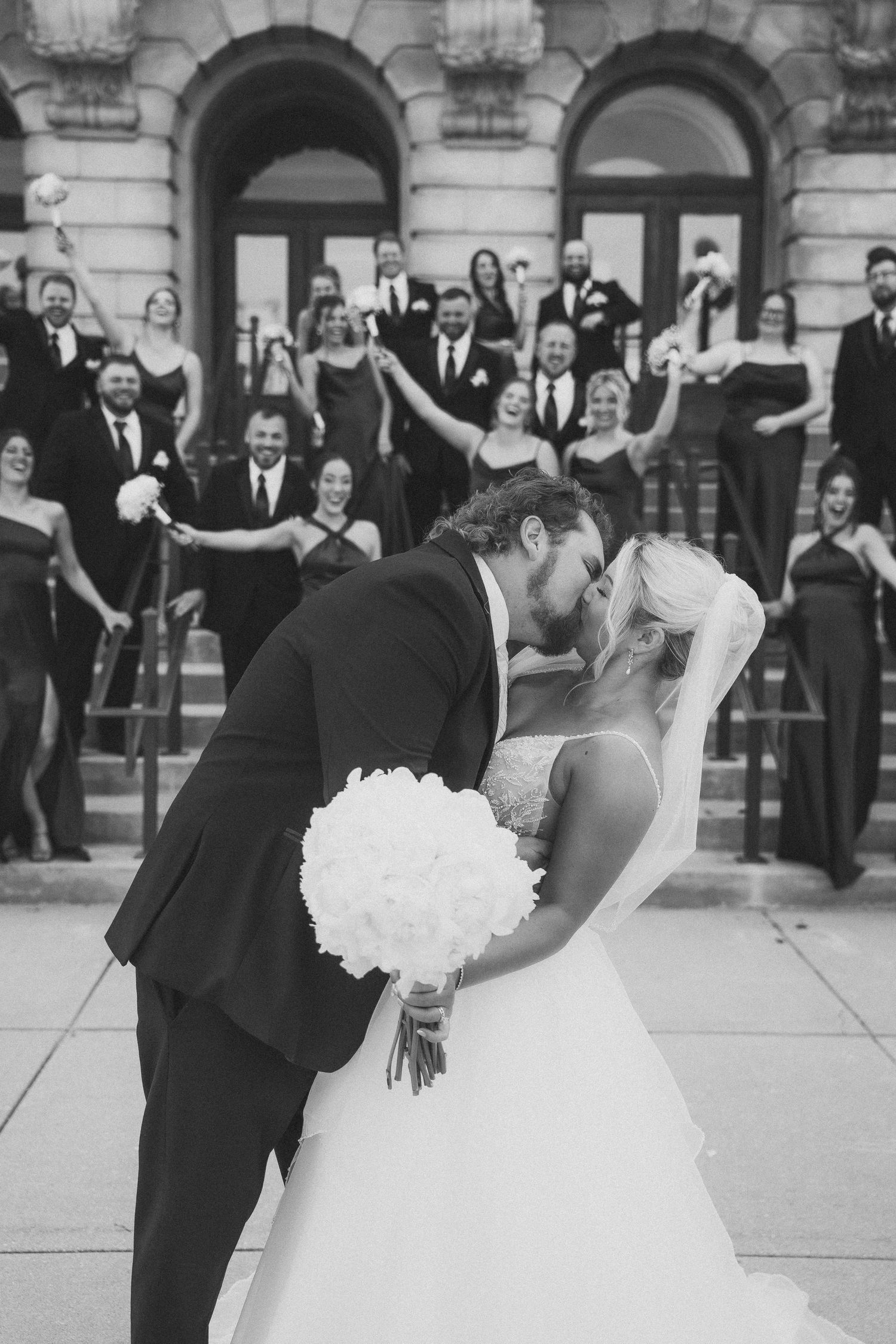 A romantic wedding couple shares a dip kiss in front of a classic building while guests look on and celebrate.