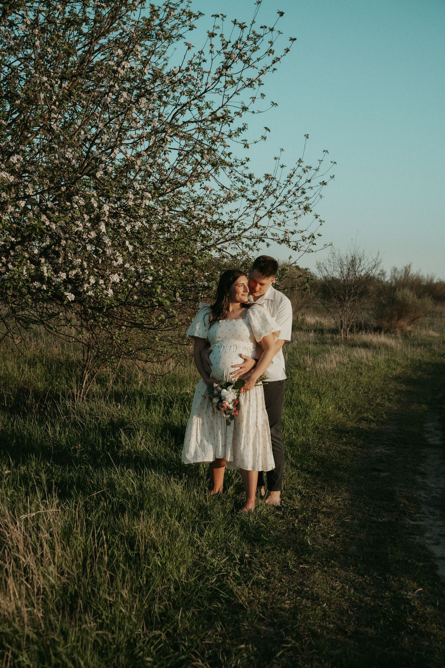 Couple embracing outdoors near blooming tree, pregnant woman in white dress holding flowers at golden hour.