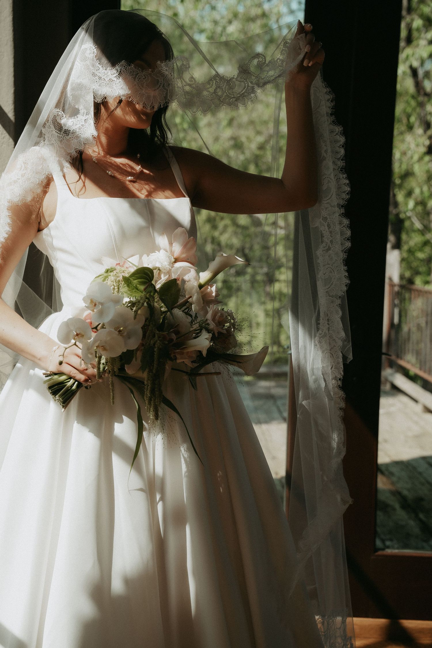 Bride in white gown and lace veil holds floral bouquet by sunlit window.
