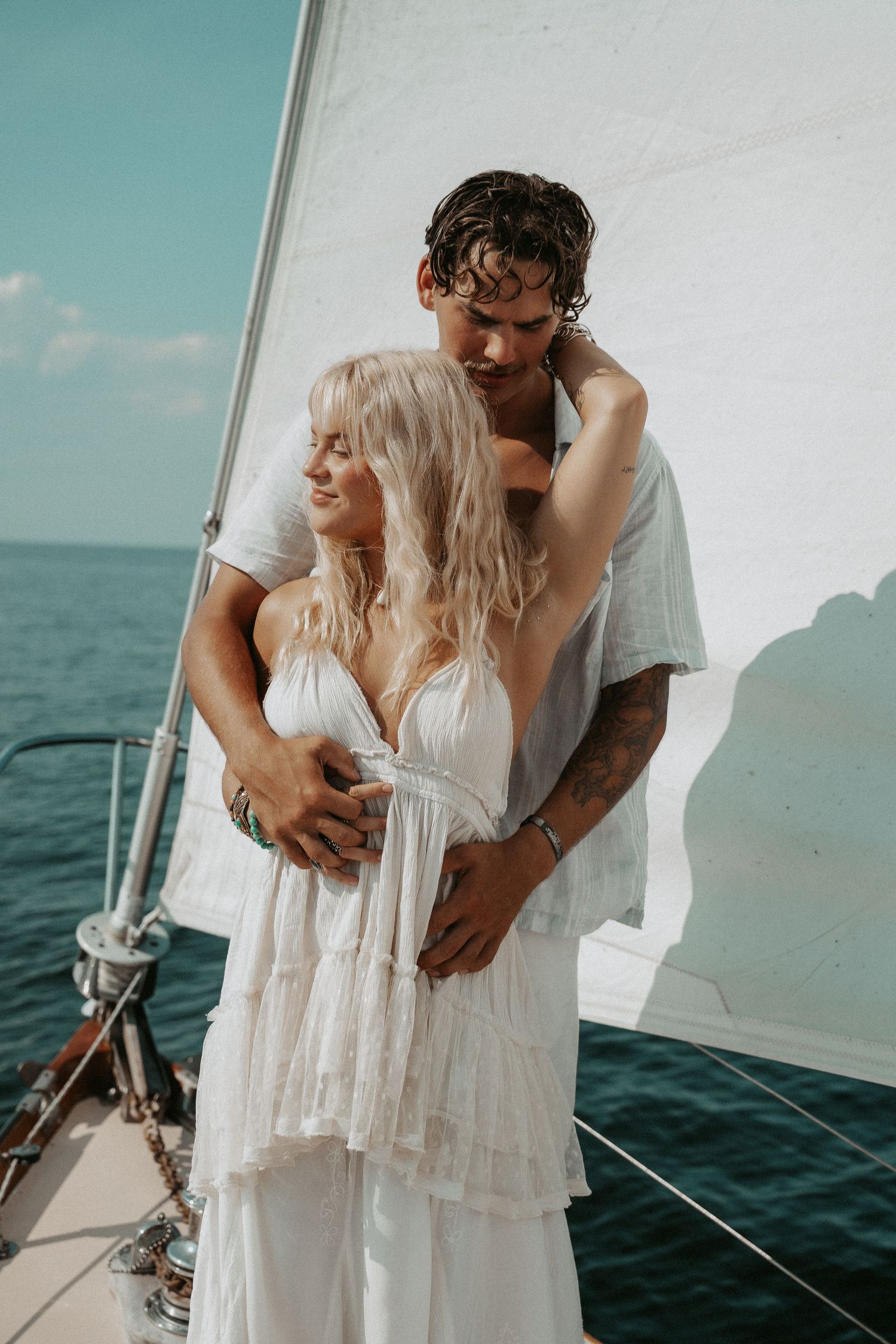 Couple embracing on a sailboat, woman in white dress, man with tattoos, ocean backdrop on a sunny day.