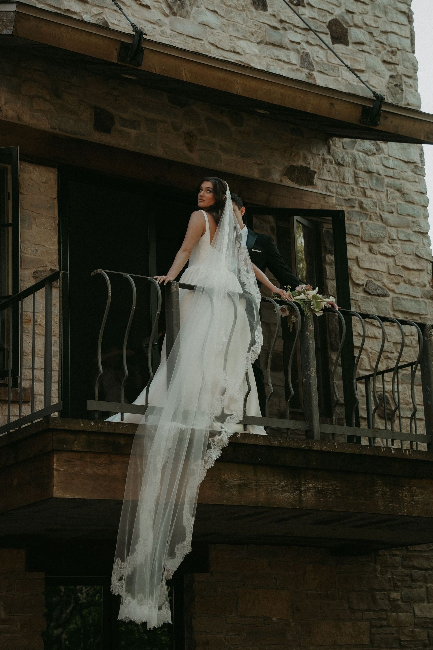 Bride in a flowing white gown and long veil stands on a stone balcony holding a bouquet on her wedding day.