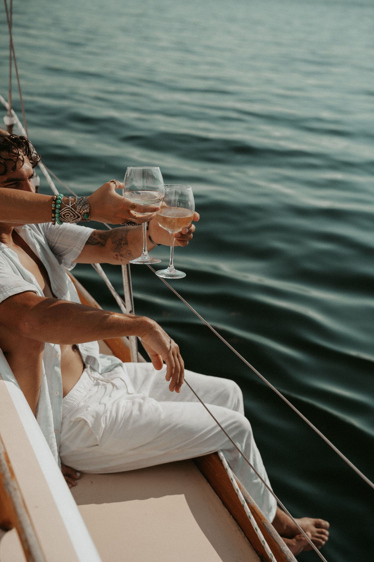 Two people toasting with wine glasses on a sailboat deck over calm turquoise water.
