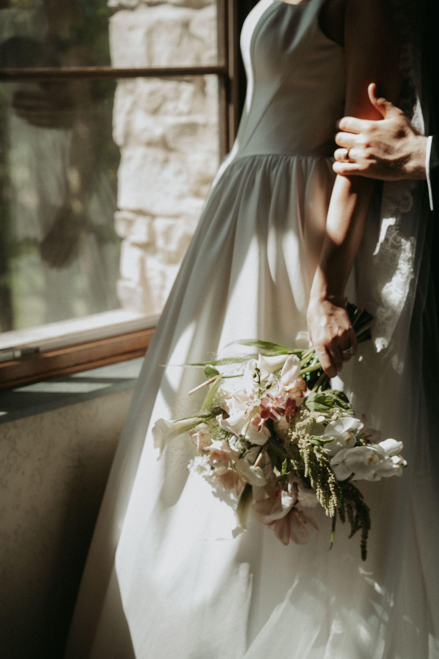 Bride in white gown holding a soft pink and white floral bouquet by a sunlit window.