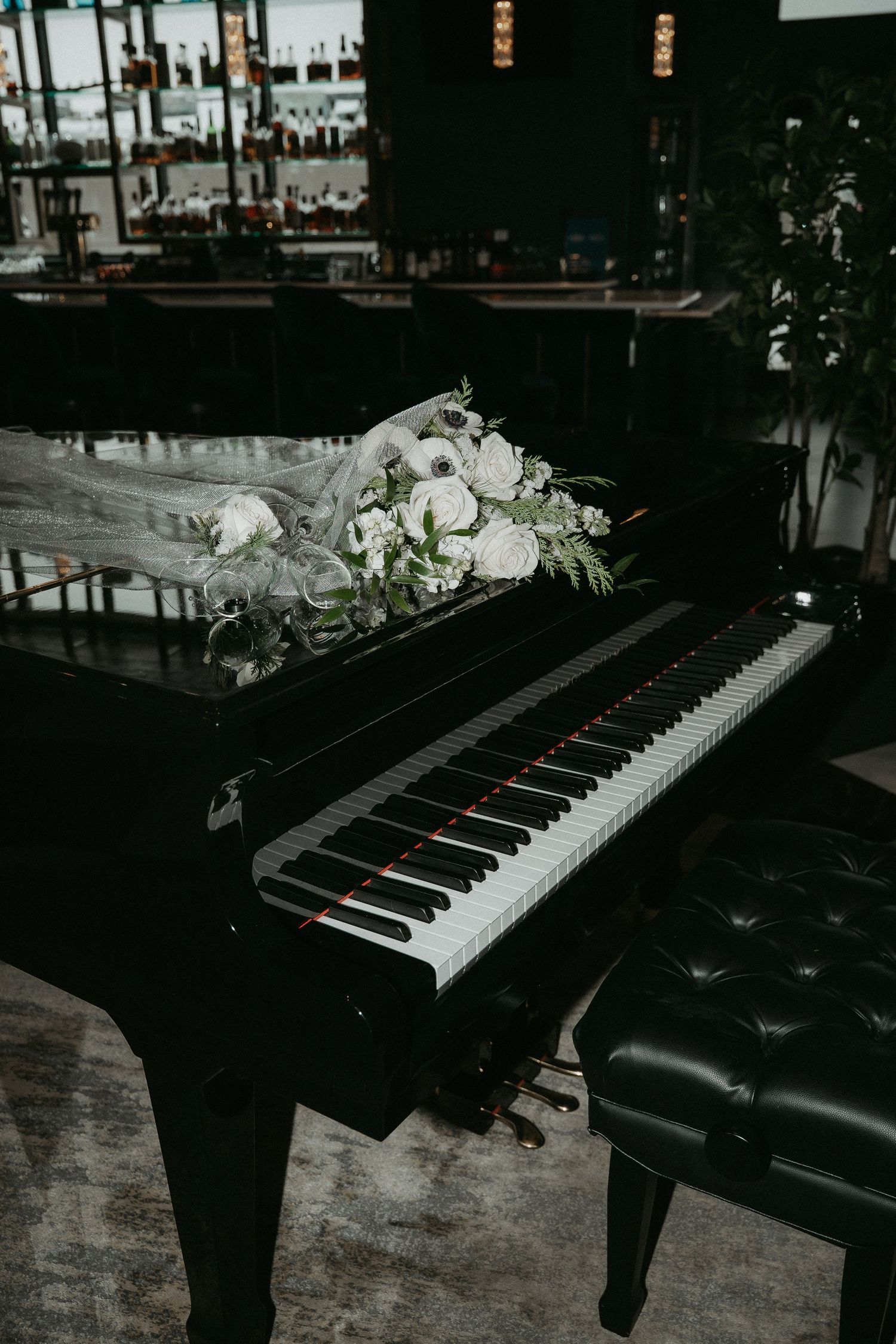 A black grand piano with white floral arrangement resting on top in a dimly lit room with a bar in background.