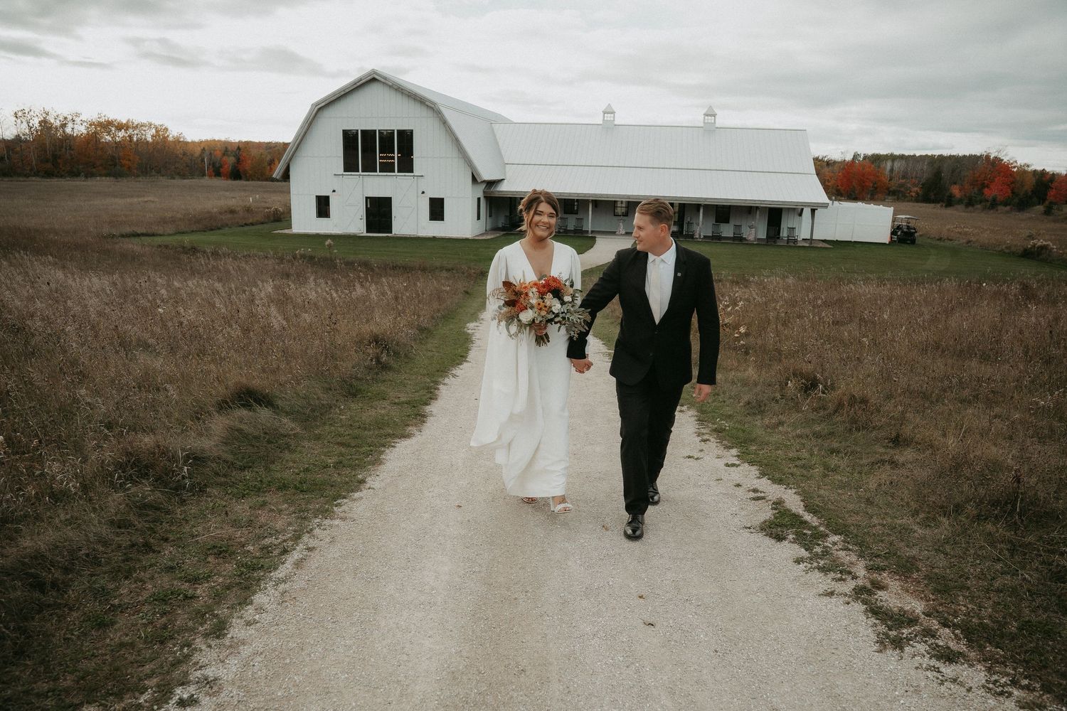 A couple walks along a rustic dirt path in an open field on their wedding day.