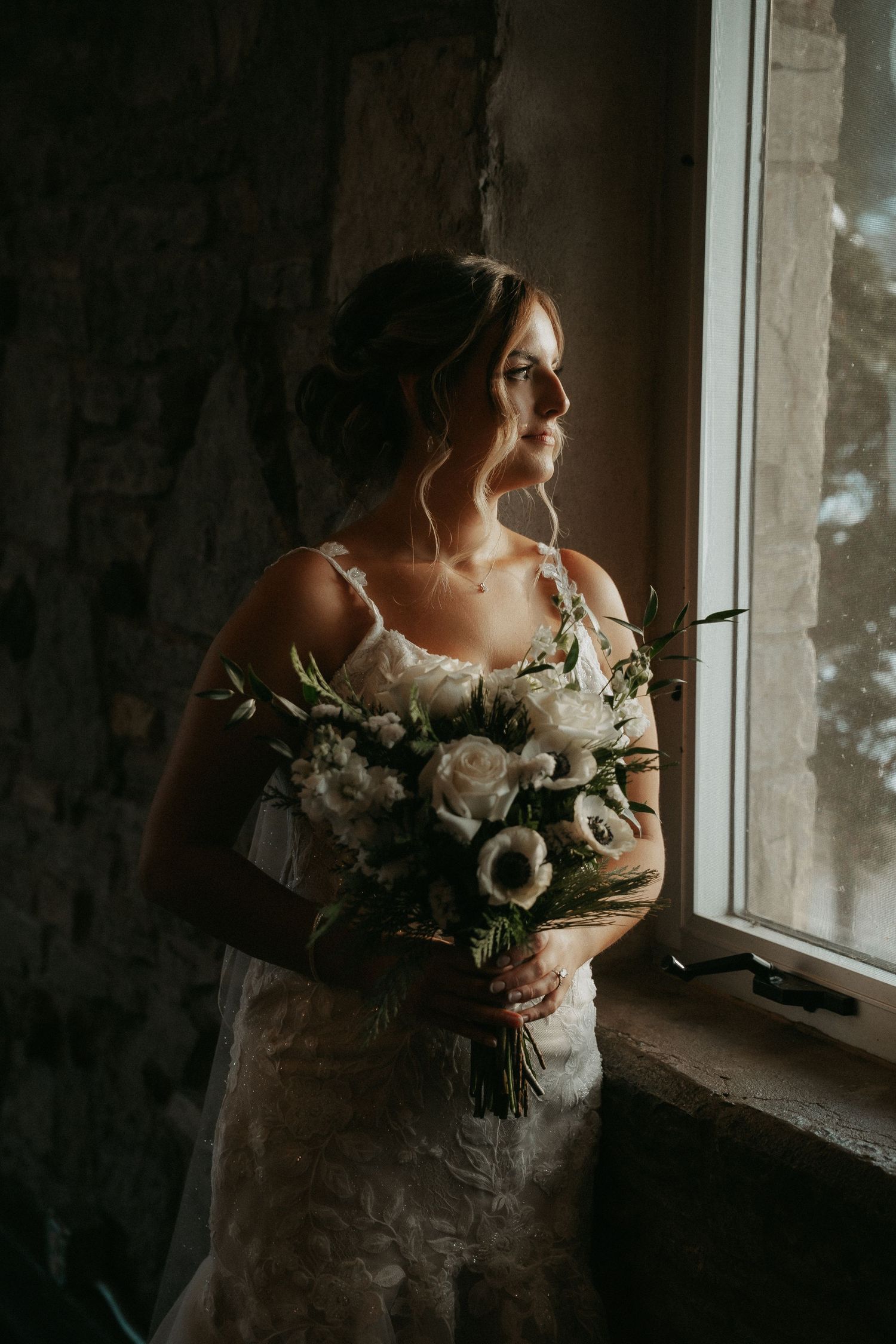 A bride in a white lace dress holds a bouquet of white flowers while gazing through a window in moody lighting.