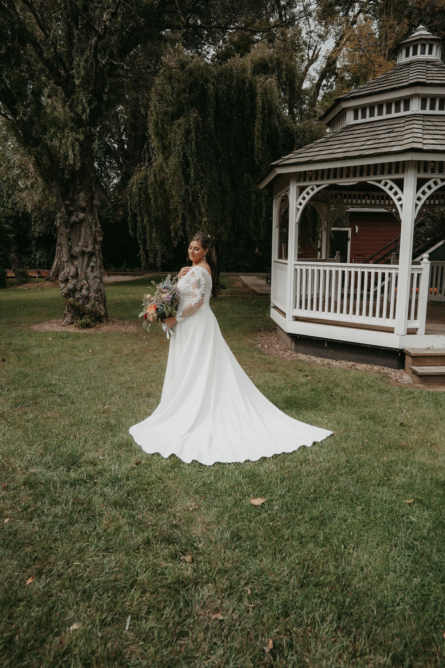 A bride in a flowing white wedding dress twirls gracefully on a lawn near a white gazebo on an autumn day.