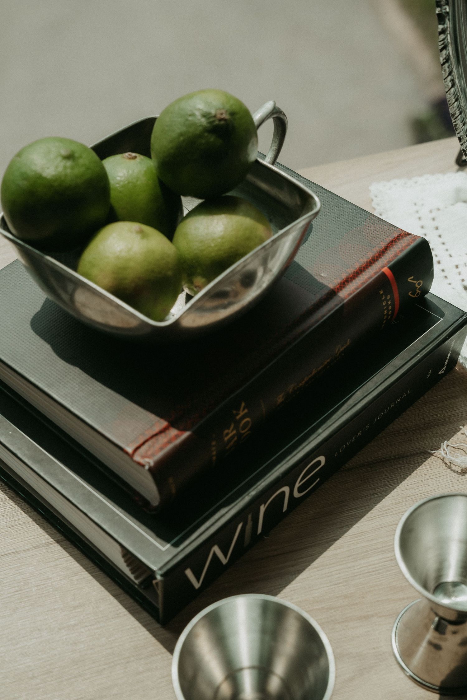 Silver bowl of fresh green limes stacked on wine books beside metal jiggers on a wooden table.
