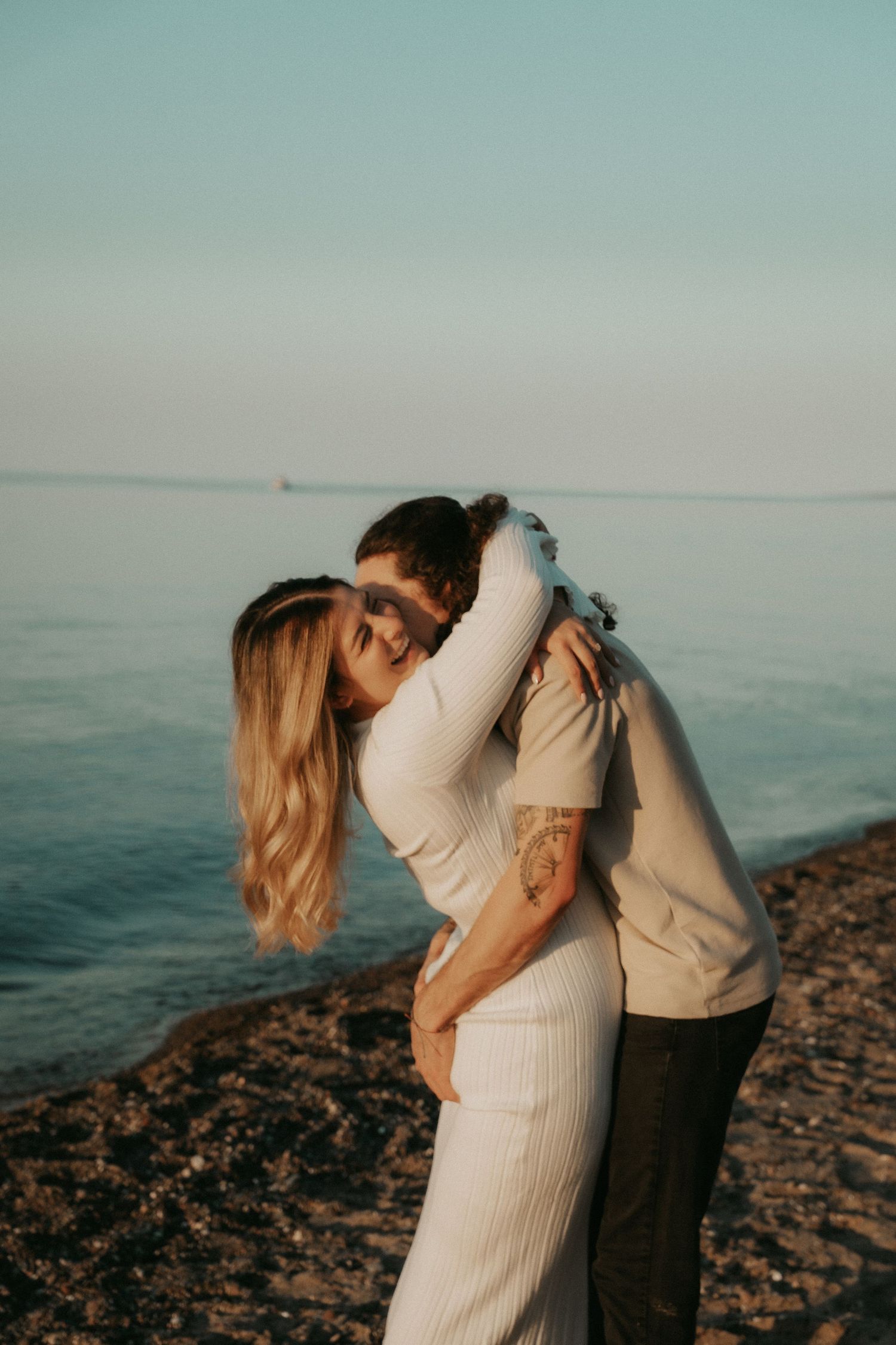 Romantic couple embraces on rocky beach at sunset with ocean in background.