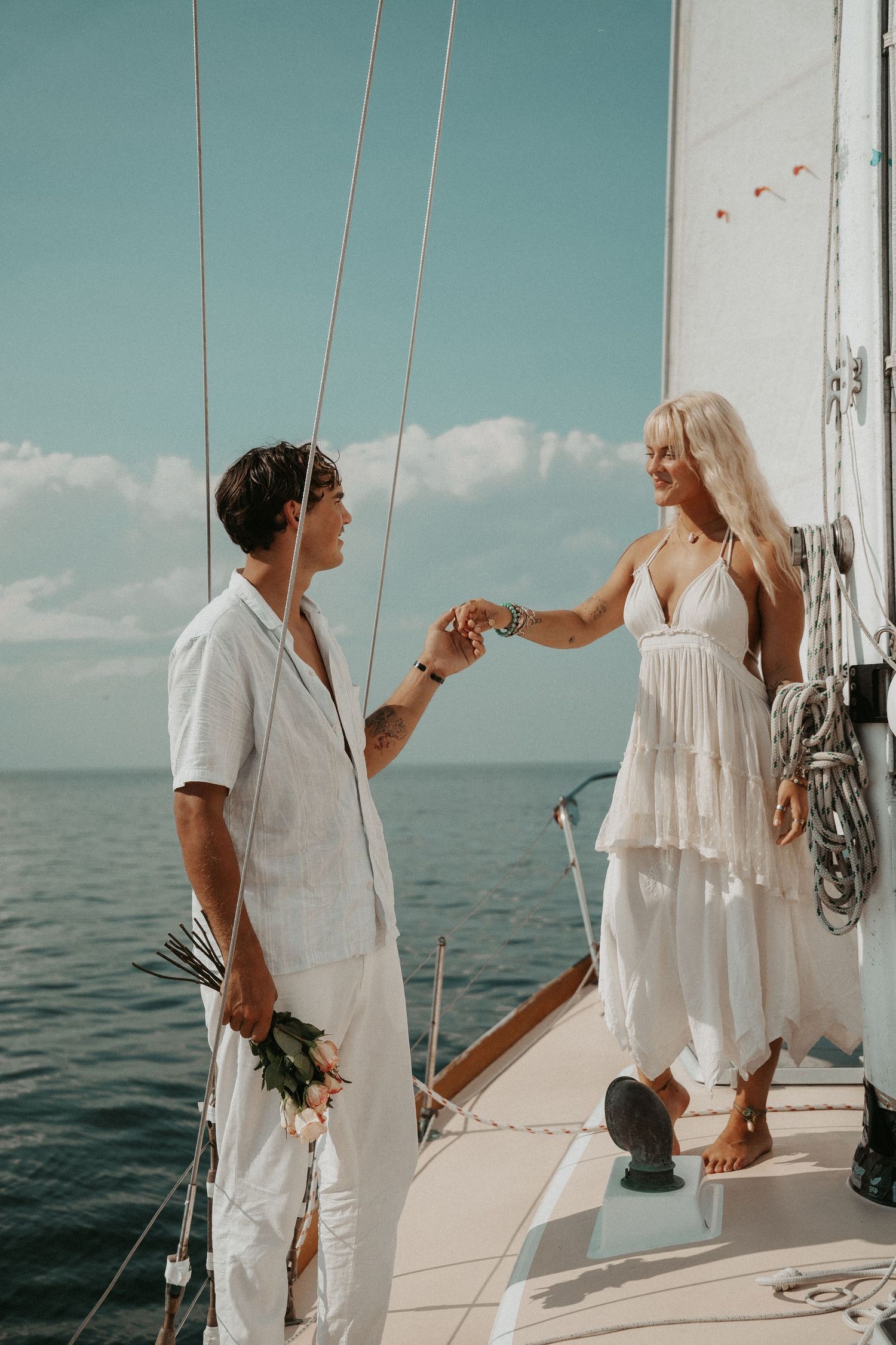 Two women in white outfits hold hands on a sailboat deck with ocean views on a sunny day.