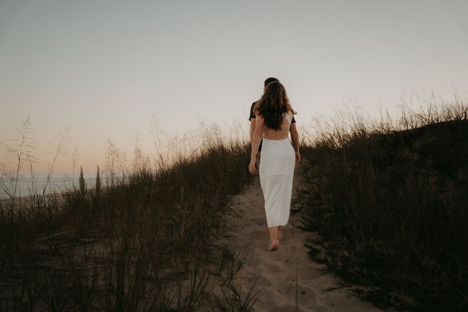 Person in white dress walks along grassy path at sunset creating silhouette against dusky sky.