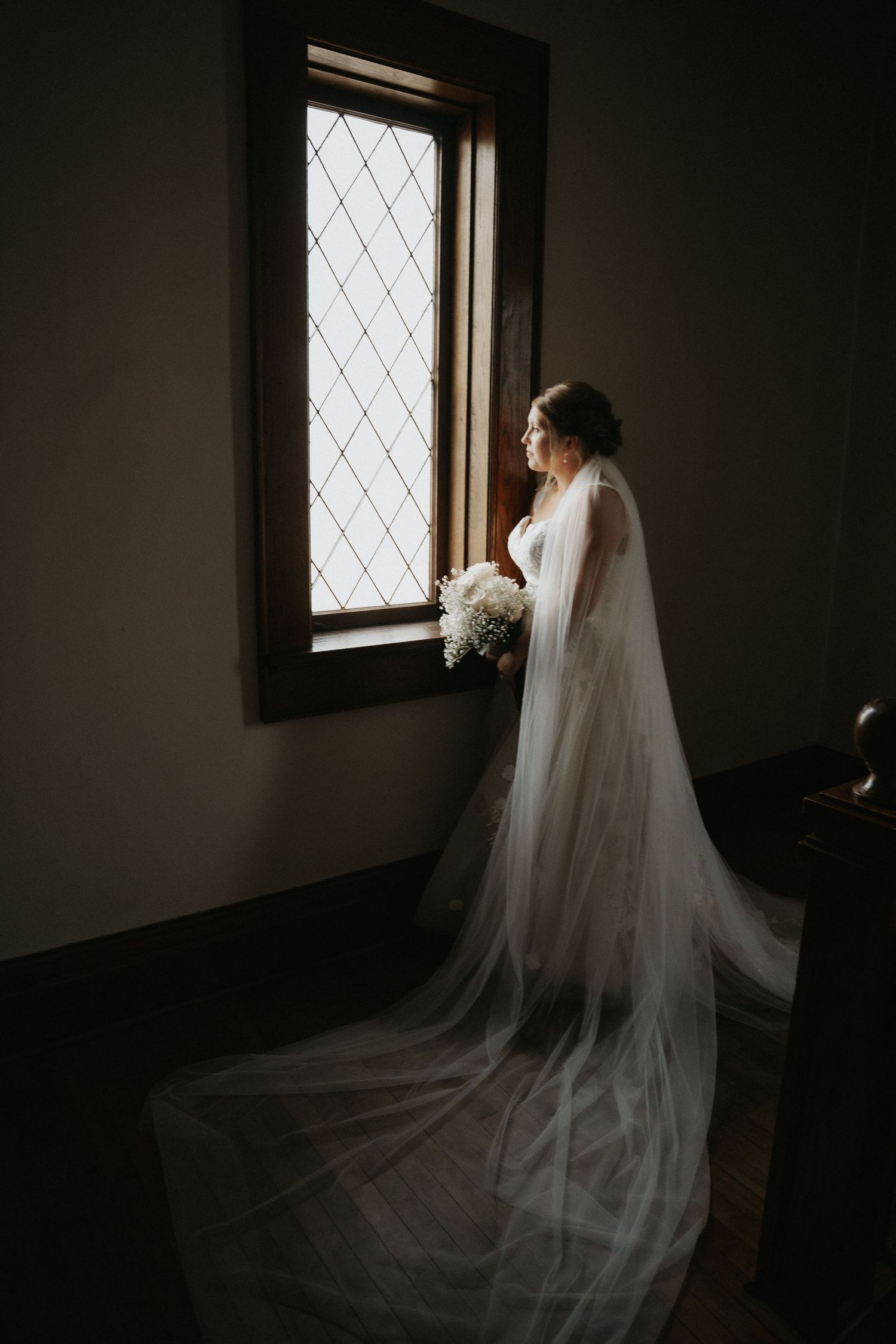 A bride in a flowing white veil stands in silhouette against a window in a dark, moody setting.