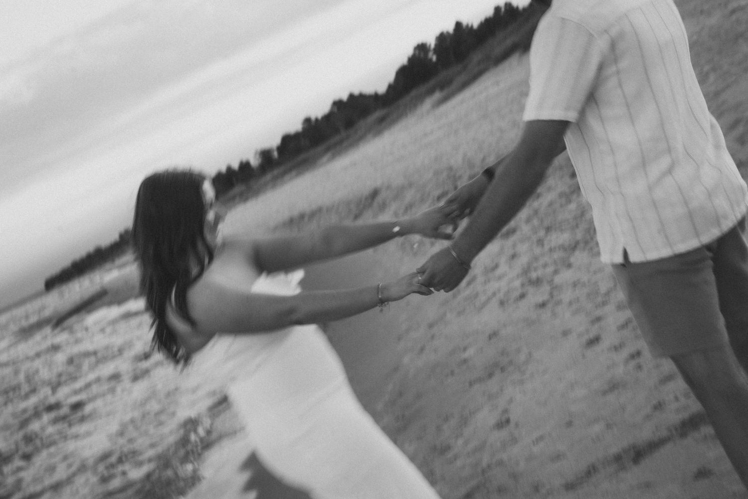 Black and white photo of two people holding hands and spinning on a beach in a playful moment.
