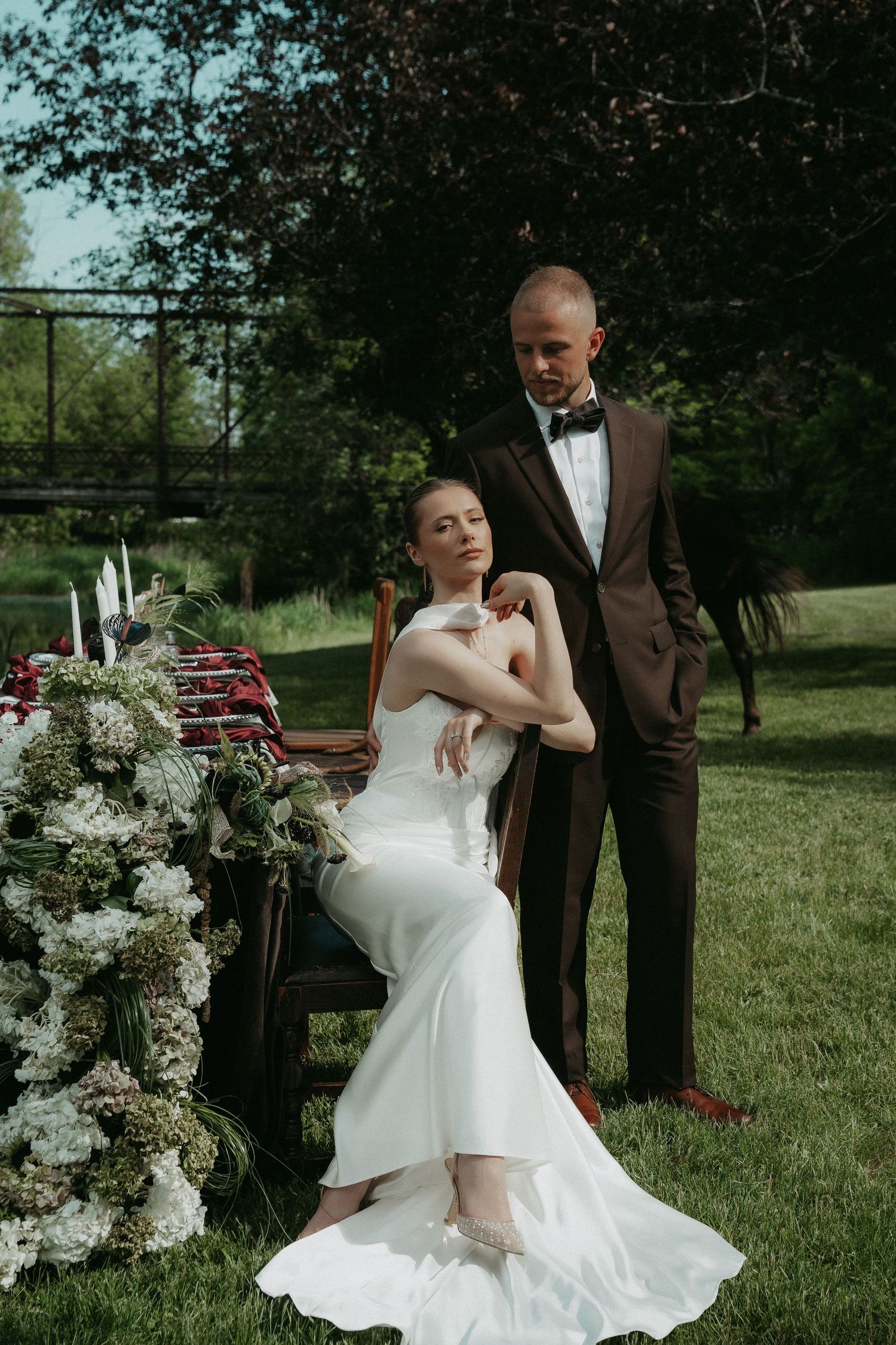 A bride in a white gown sits beside a floral arrangement while a groom in a brown suit stands beside her outdoors.