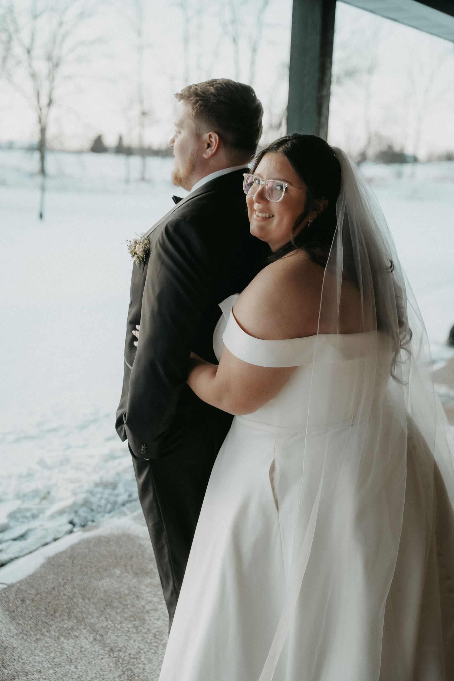 A bride in a white dress and veil stands behind her groom during their first look moment in a snowy winter setting.
