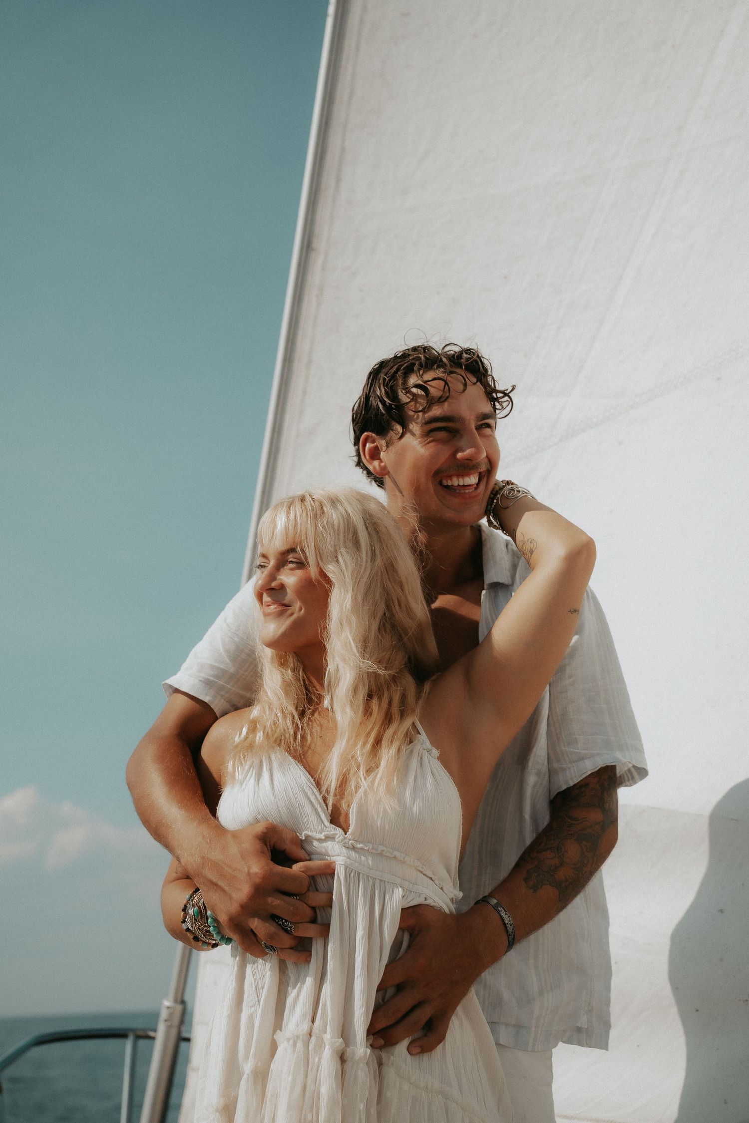 A smiling couple embraces on a sailboat, wearing white outfits, with the sail and blue sky behind them.