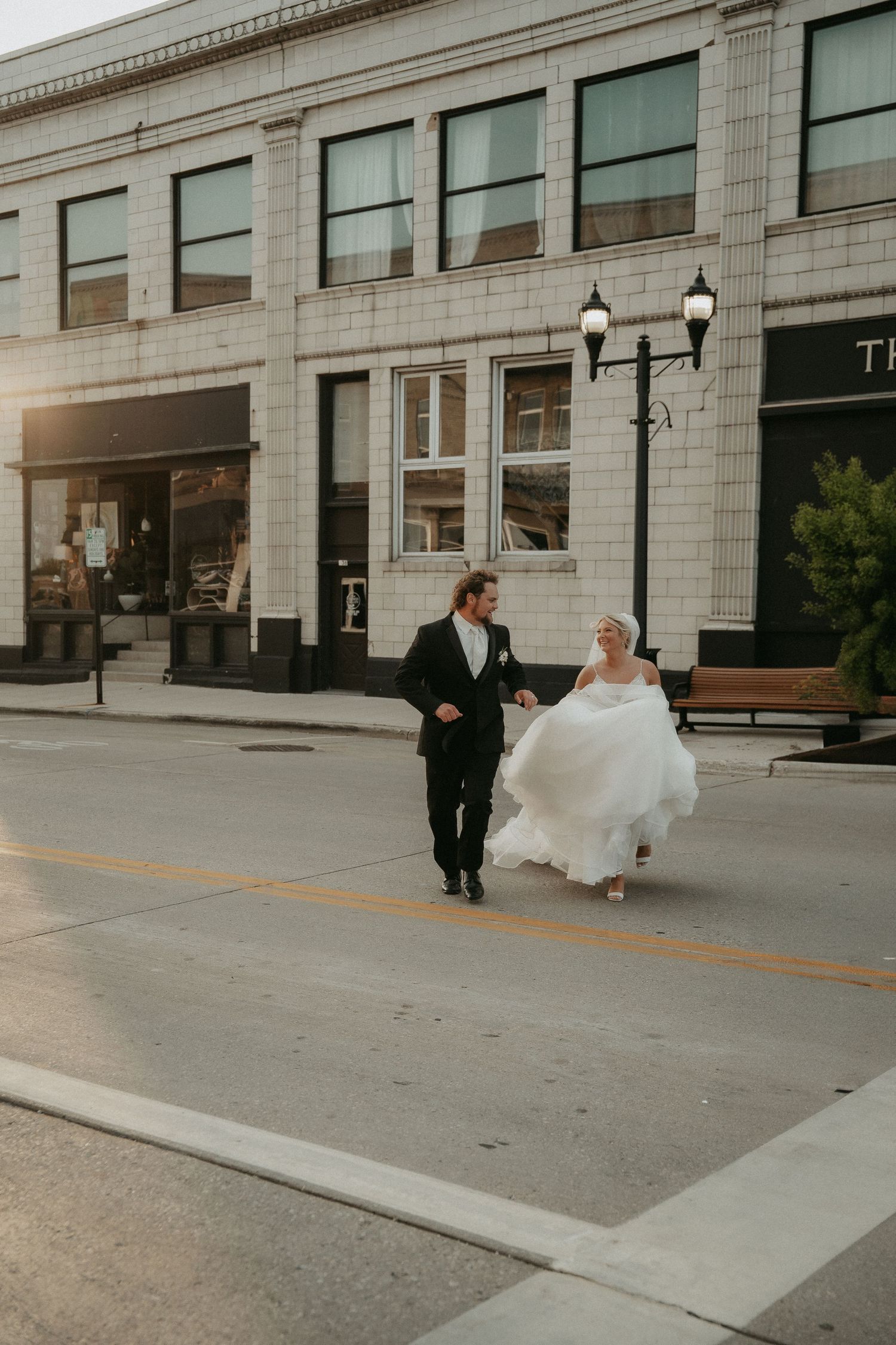 A wedding couple runs playfully down a city sidewalk with classic stone architecture and lamp posts in the background.