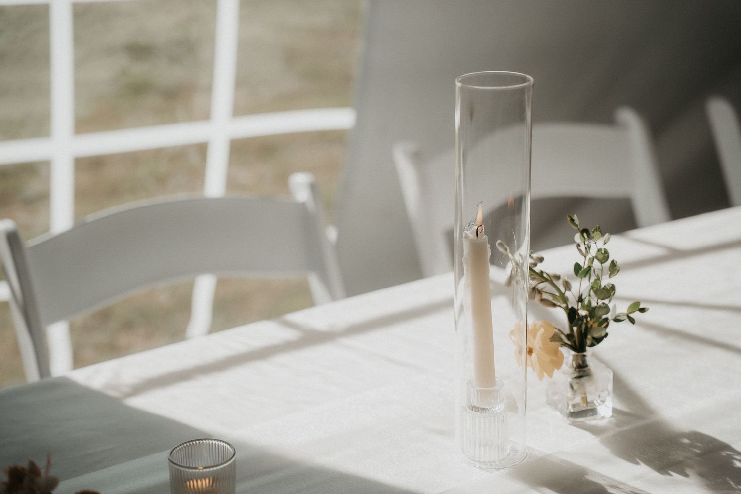 A minimalist white wedding reception table setting with delicate greenery and candles in glass holders.