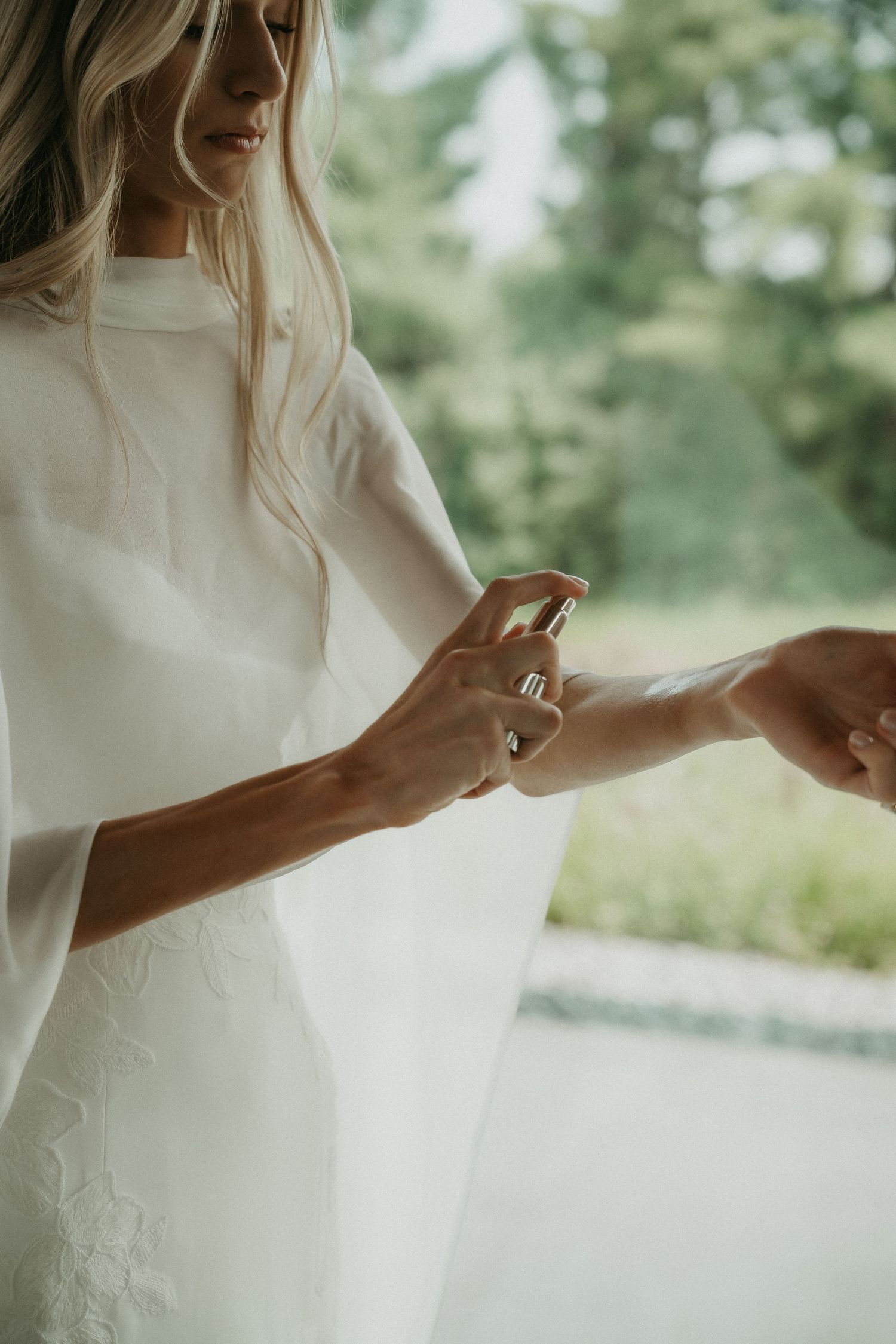 Bride in white dress spraying perfume on her wrist outdoors with lush green trees in background.