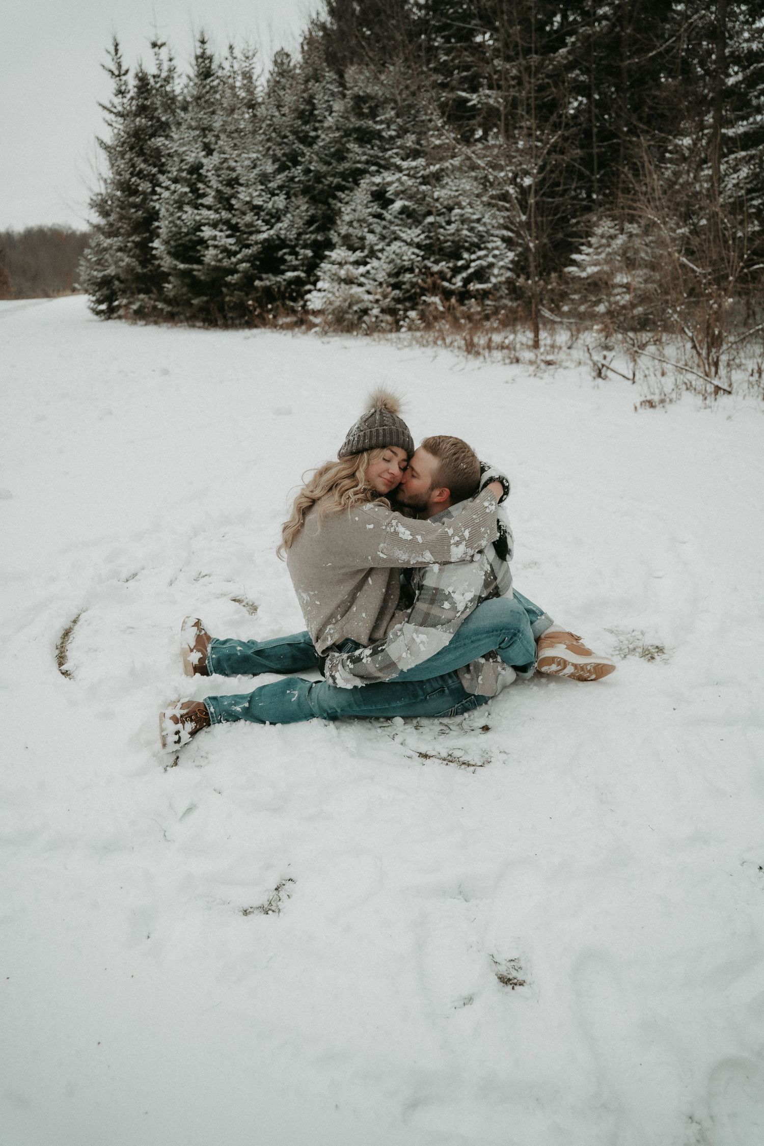 Two people in winter clothing embrace while sitting in the snow with evergreen trees in the background.