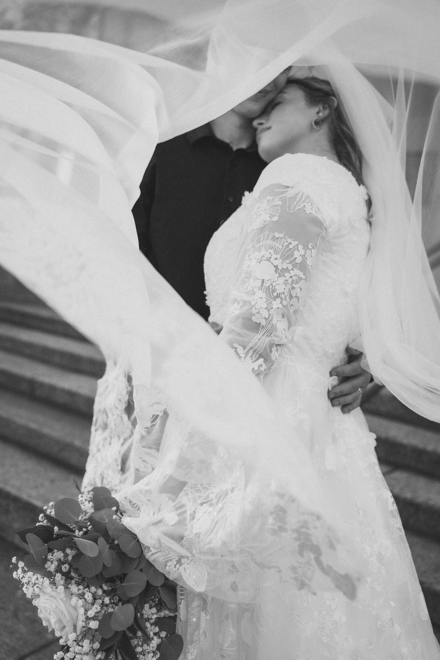 Romantic black and white portrait of newlyweds embracing under flowing veil.