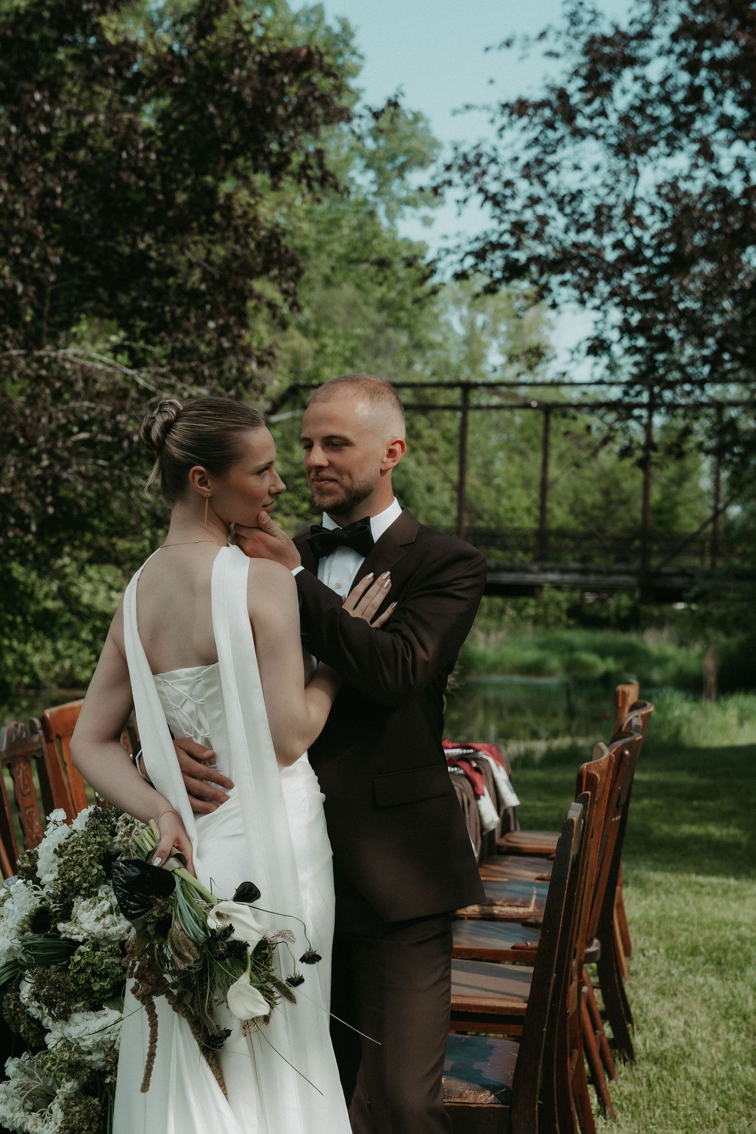 Bride and groom embrace outdoors near wooden chairs and a bridge, she holds a white floral bouquet in a sleek wedding gown.