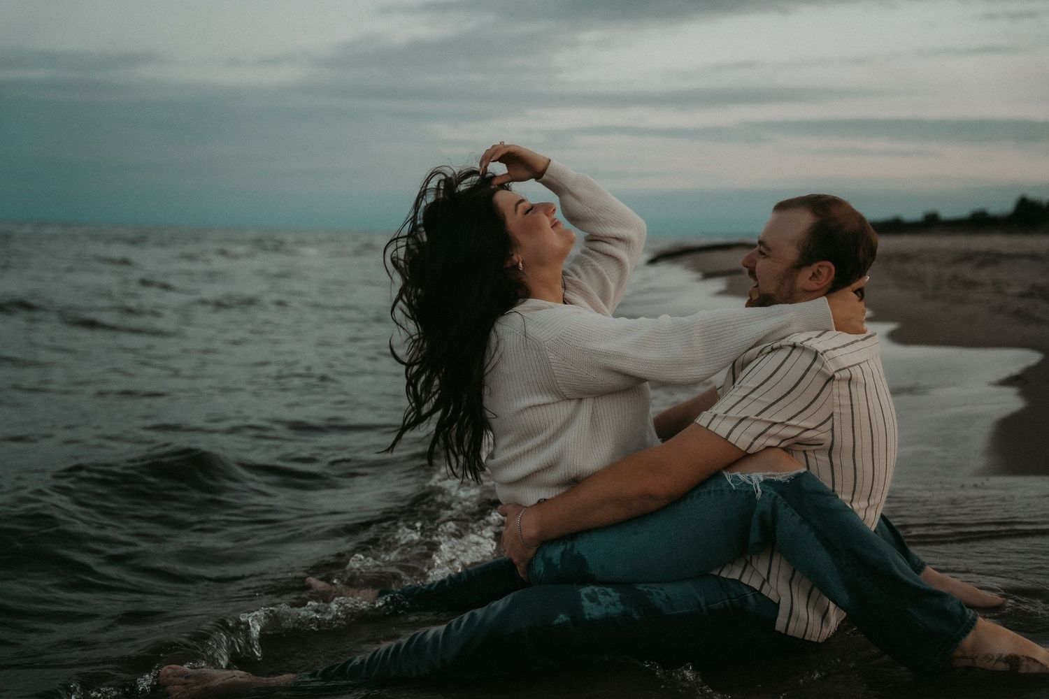 A couple shares a joyful moment together on a rocky beach at sunset with dark waves crashing nearby.