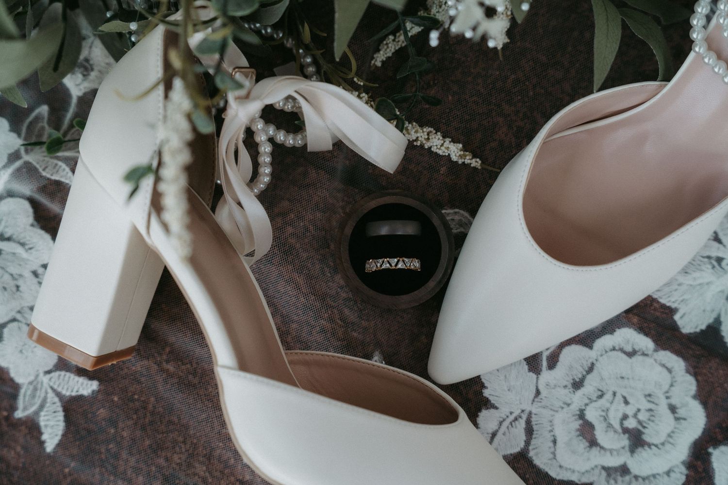 Wedding details featuring white bridal shoes, delicate floral arrangements, and elegant accessories on a dark surface.