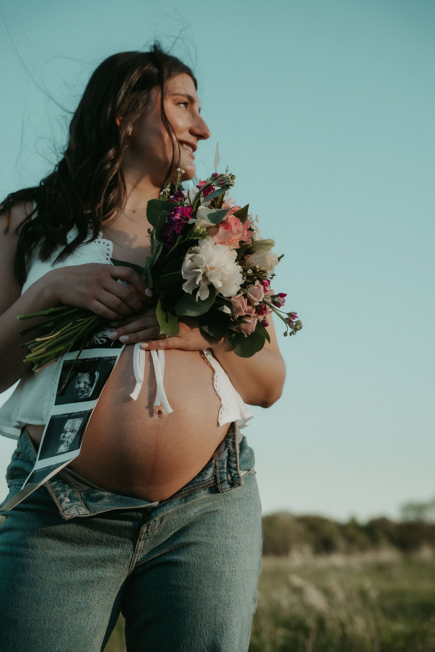 Smiling pregnant woman holding a colorful flower bouquet outdoors, with ultrasound photos tucked in her jeans waistband.