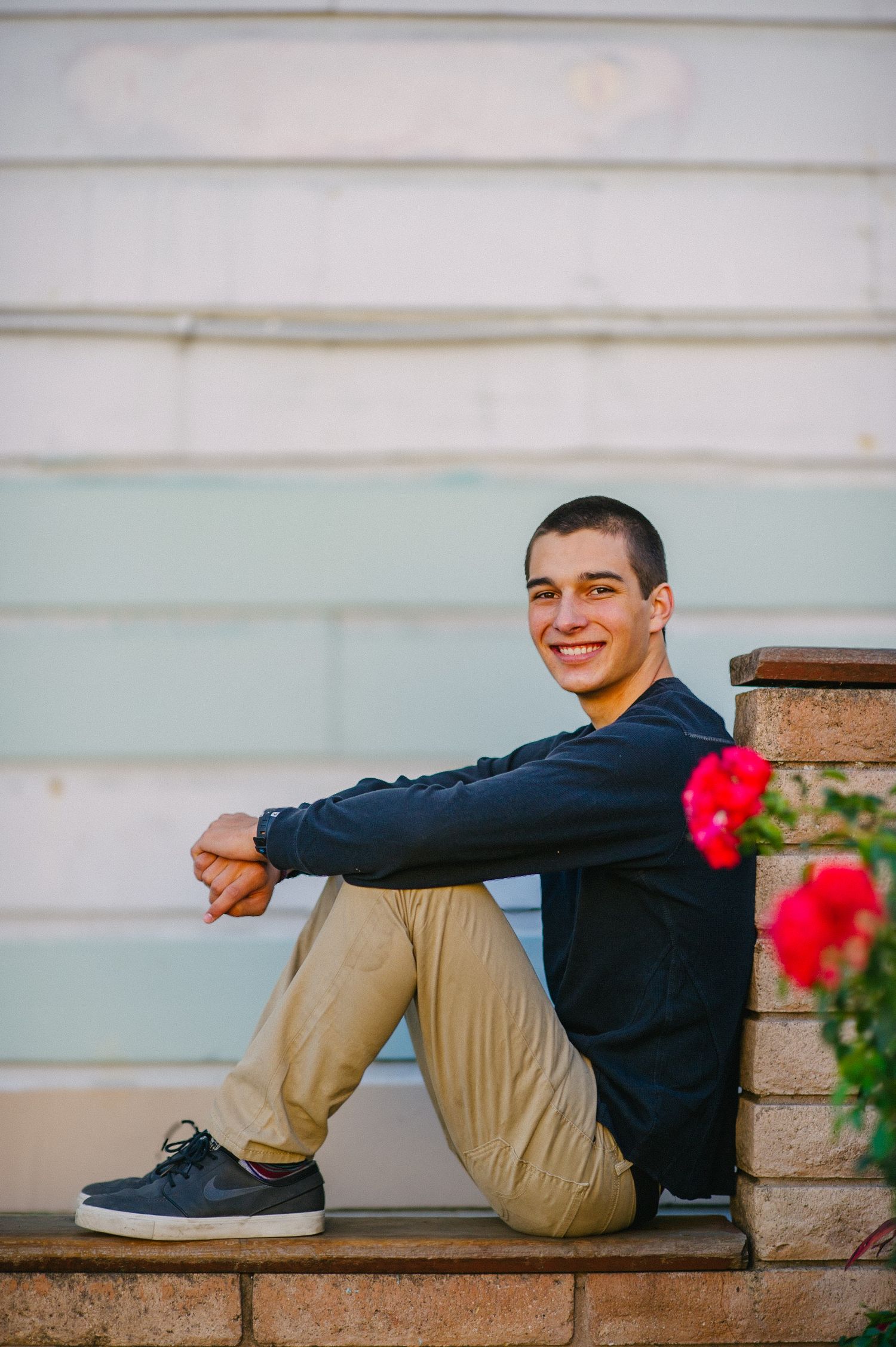 Person sitting on concrete steps wearing navy jacket and khaki pants with red flowers in foreground.