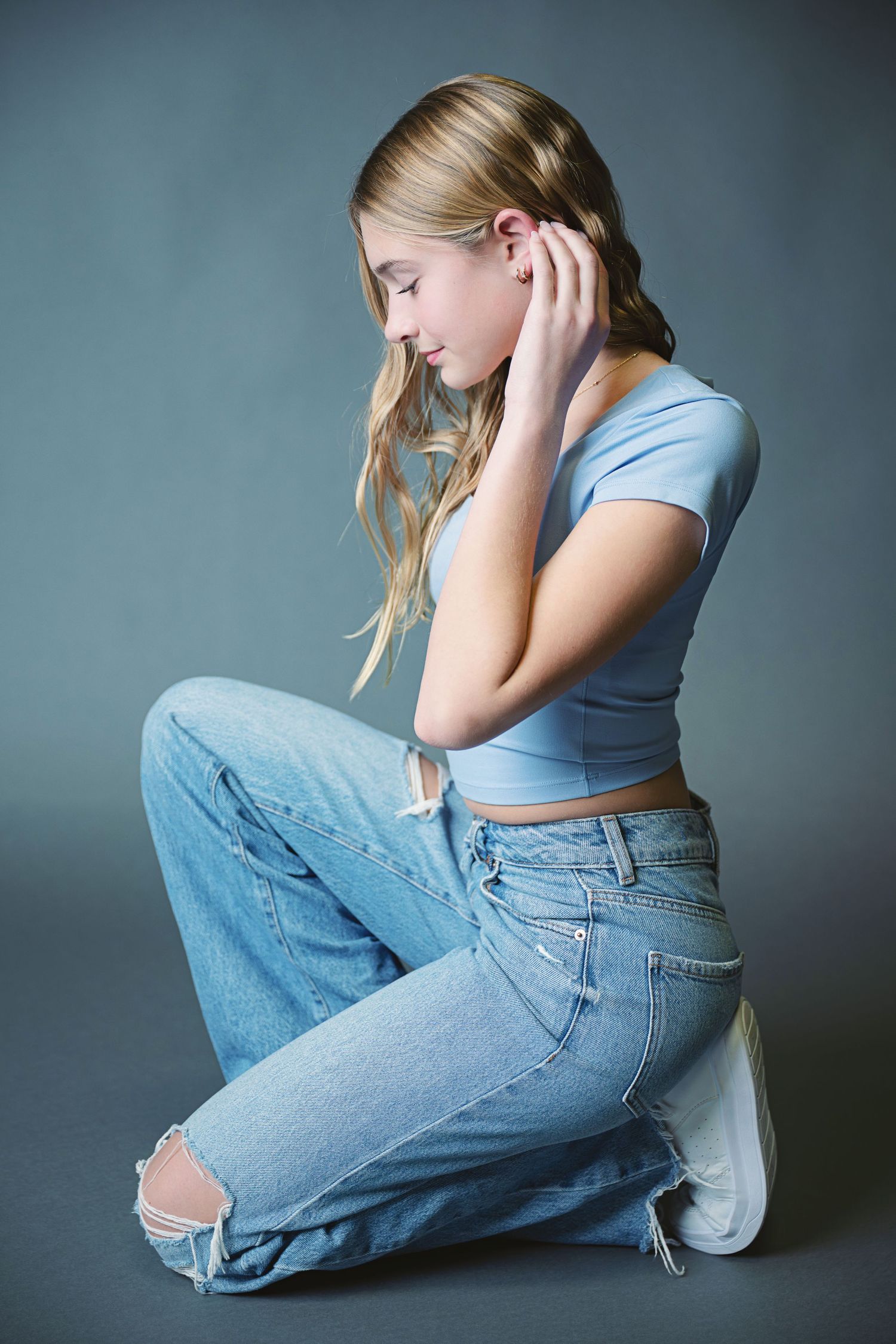 Person in light blue top and distressed jeans sits in contemplative poses against gray studio backdrop.