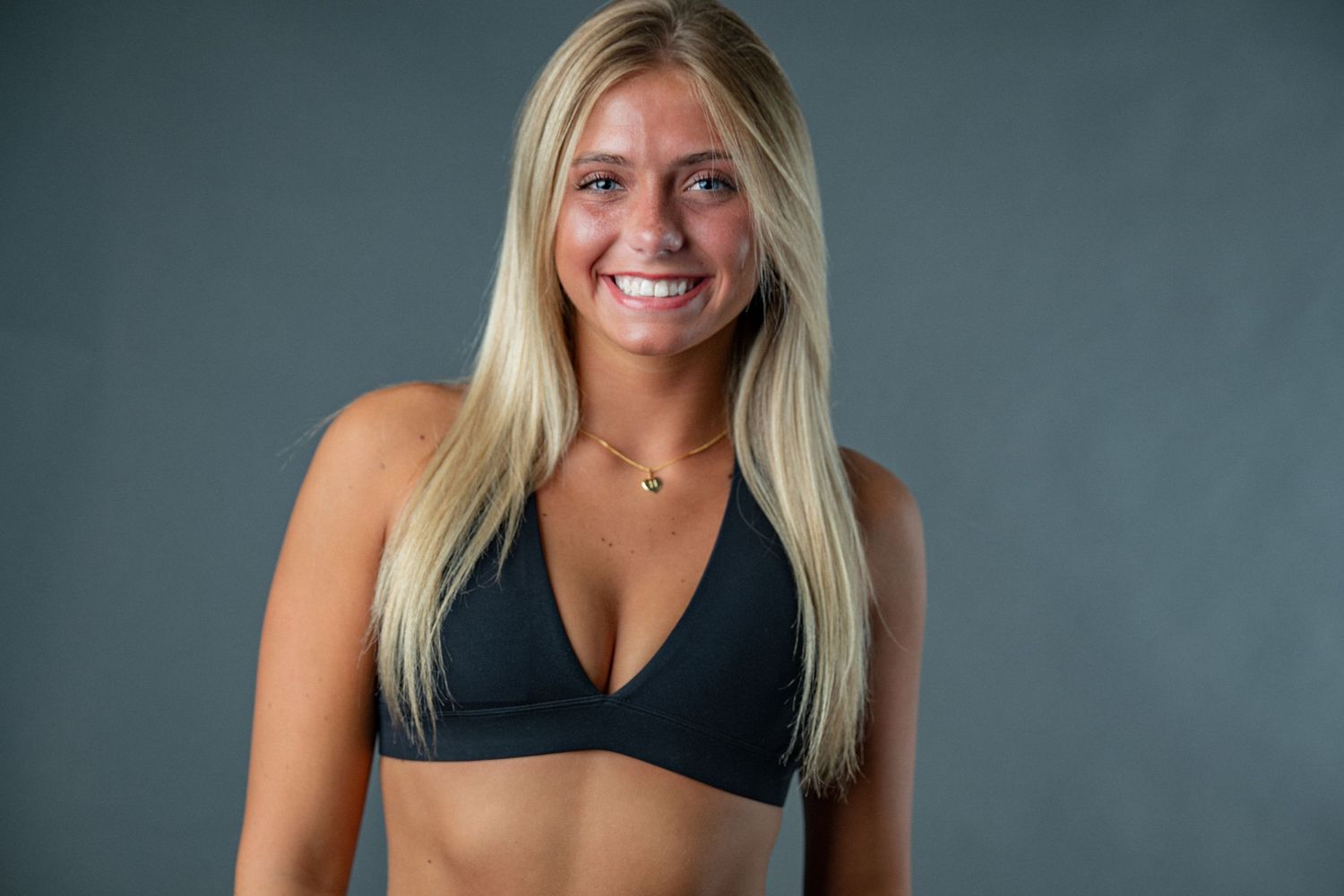 Athletic woman with long blonde hair wearing a black sports bra poses against gray studio backdrop.