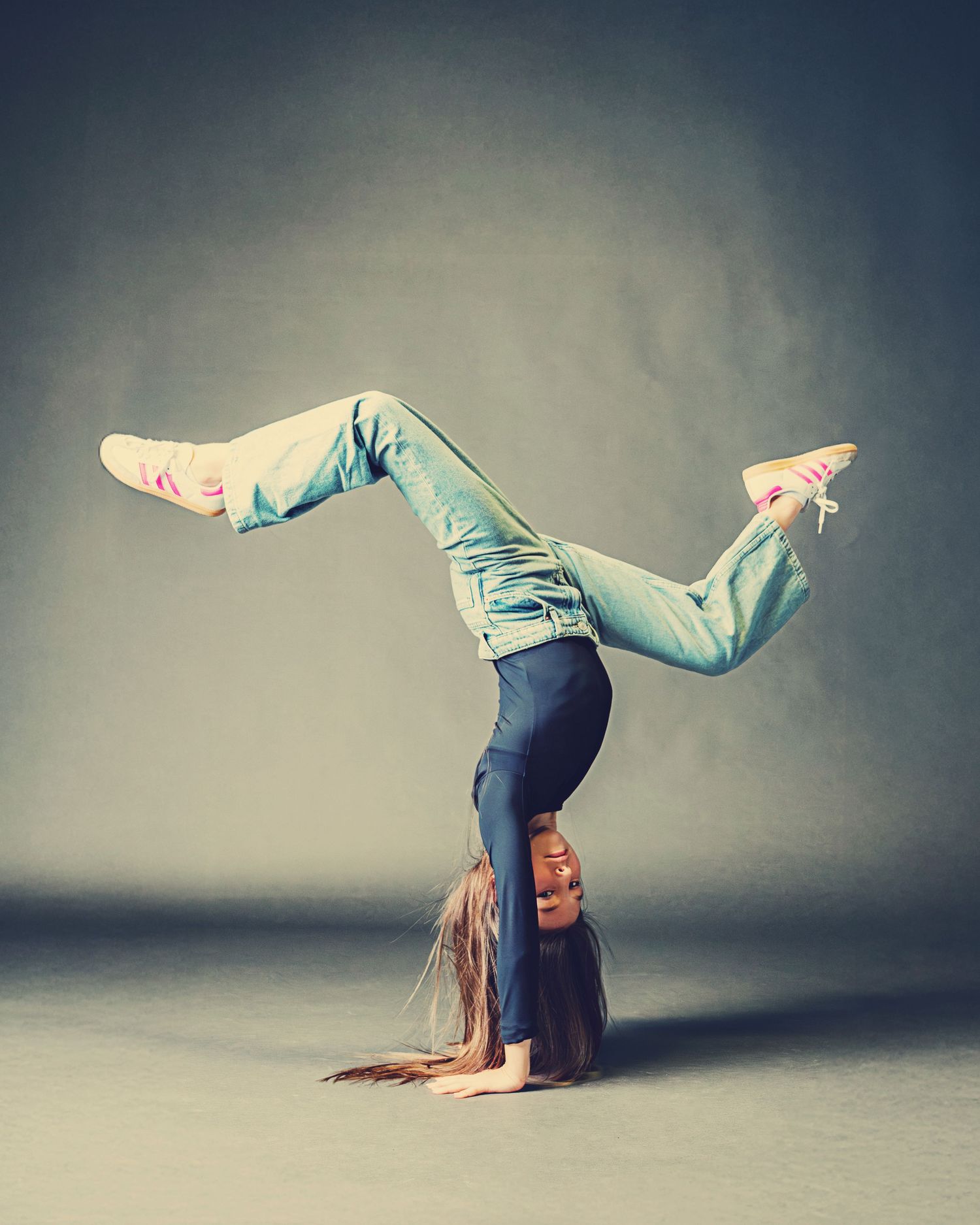 A dancer performs a handstand split wearing light blue jeans and a dark top against a muted gray background.
