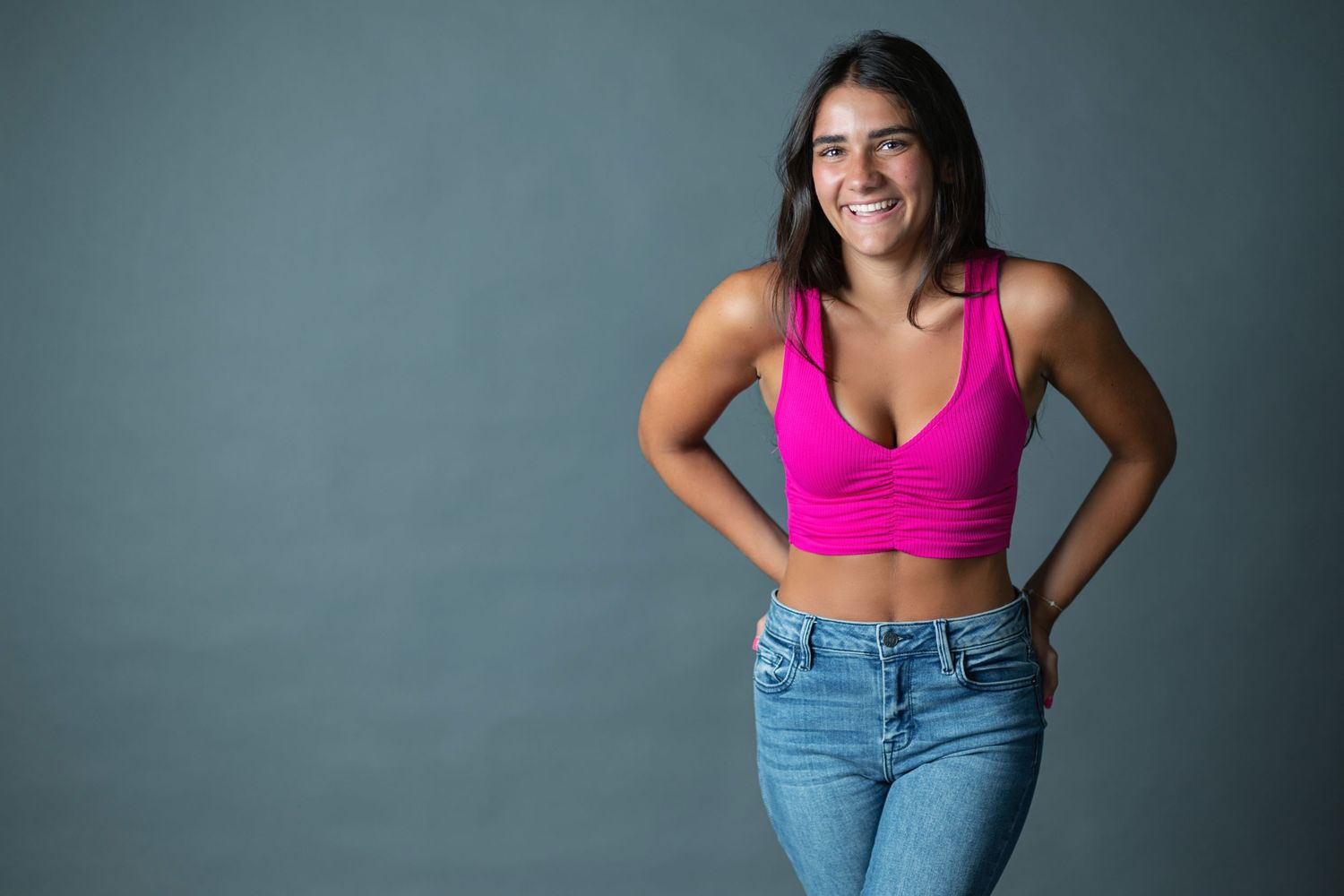 A person in a pink sports bra and blue jeans poses confidently against a gray background.