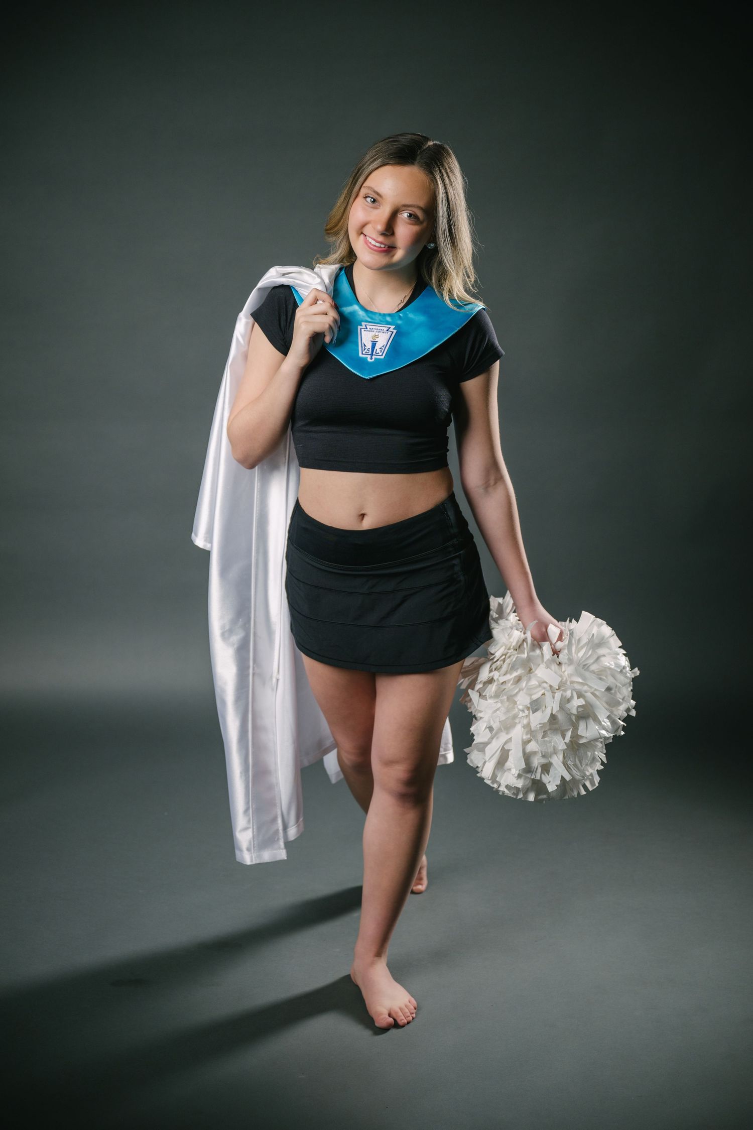 A cheerleader in black athletic wear holds white pom poms while posing against a dark gray studio backdrop.