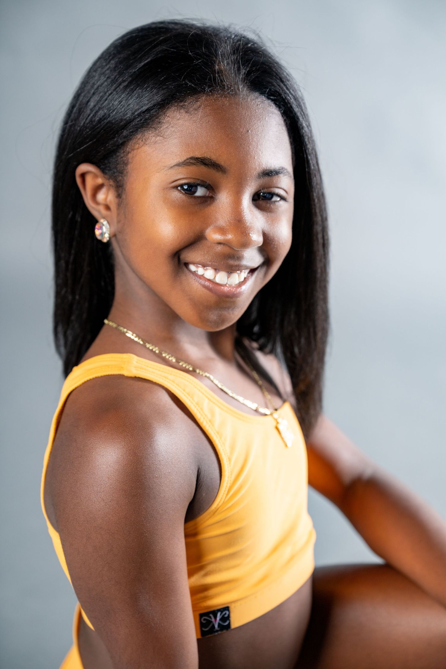 Athletic woman in yellow sports bra smiles warmly during fitness photoshoot against gray background.