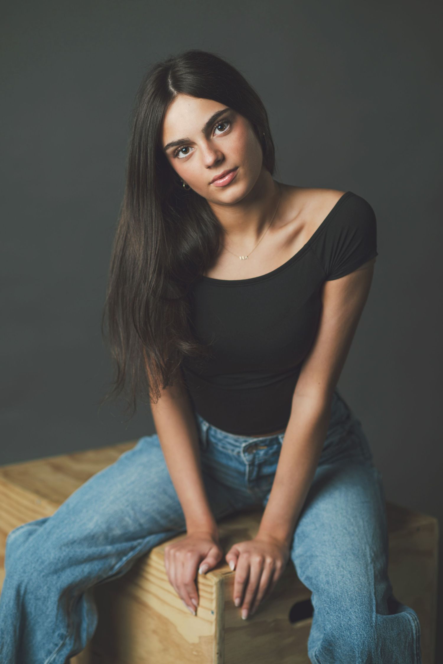 A model in a black top and blue jeans poses casually on a wooden surface against a dark gray backdrop.