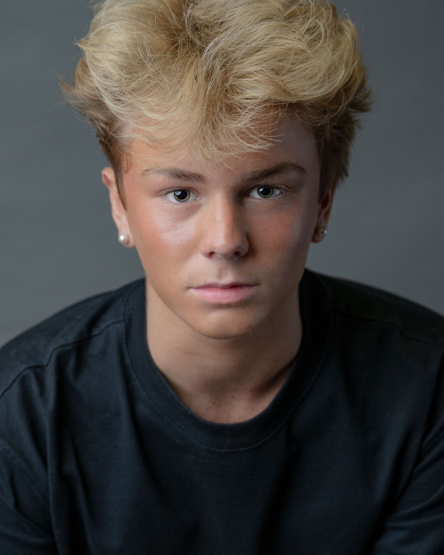Portrait of a young person with blonde spiked hair wearing a black t-shirt against a gray background.