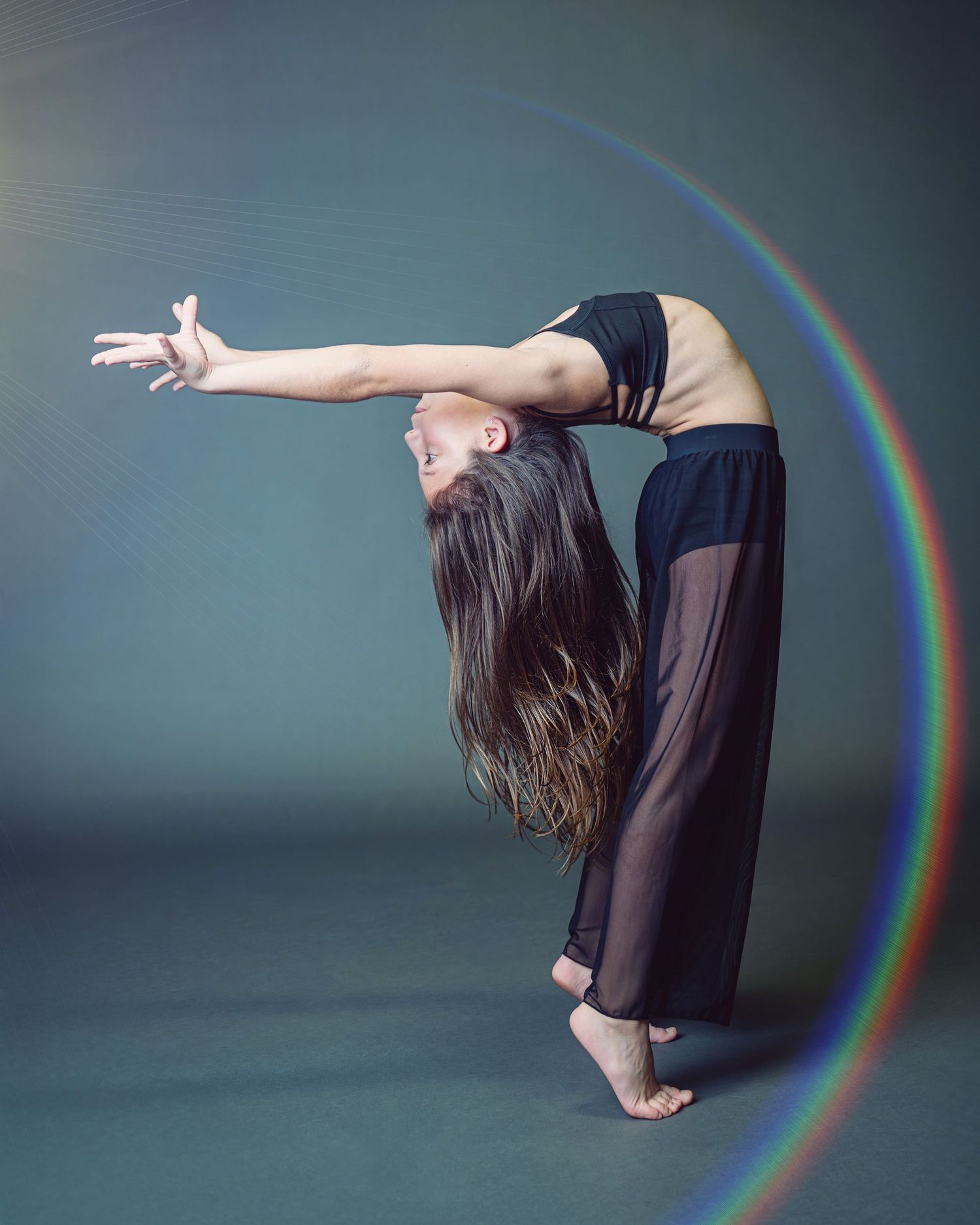 A dancer performs a graceful backbend pose against a dark background with a rainbow light effect in the frame.